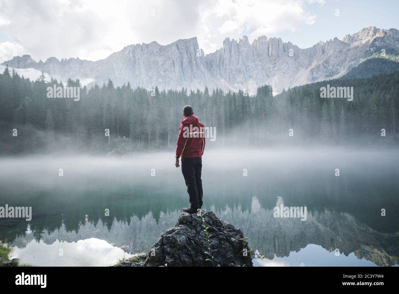 Italien, Karersee, junger Mann steht auf dem Felsen und schaut bei Sonnenaufgang auf den Lago di Karersee in den Dolomitenalpen Stockfoto