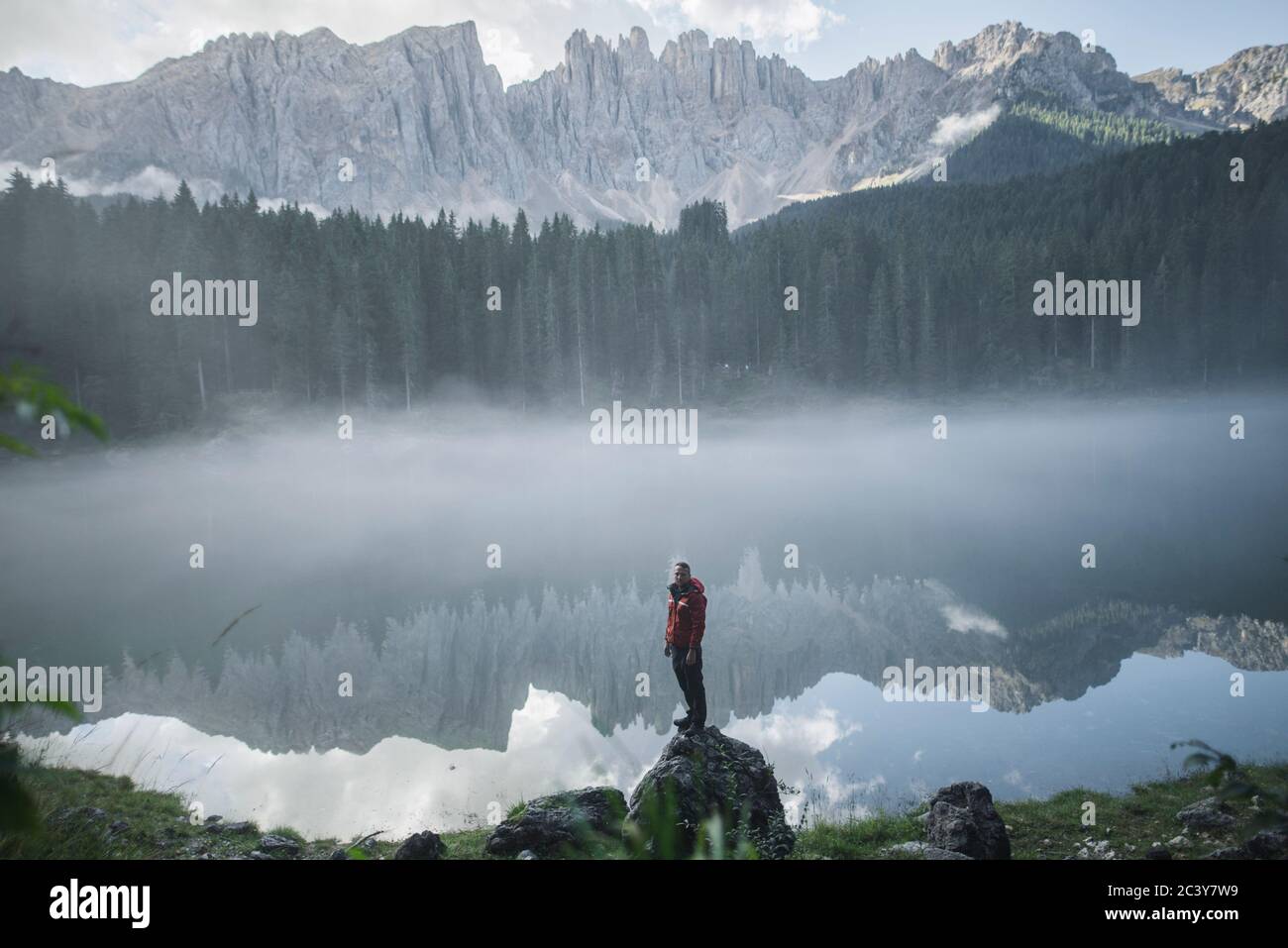 Italien, Karersee, junger Mann steht am Karersee in den Dolomiten bei Sonnenaufgang auf dem Felsen Stockfoto