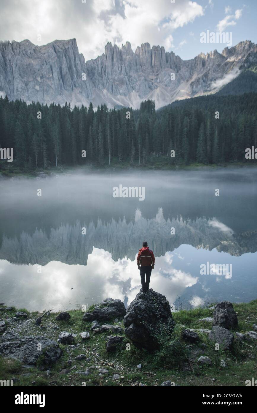 Italien, Karersee, junger Mann steht am Karersee in den Dolomiten bei Sonnenaufgang auf dem Felsen Stockfoto