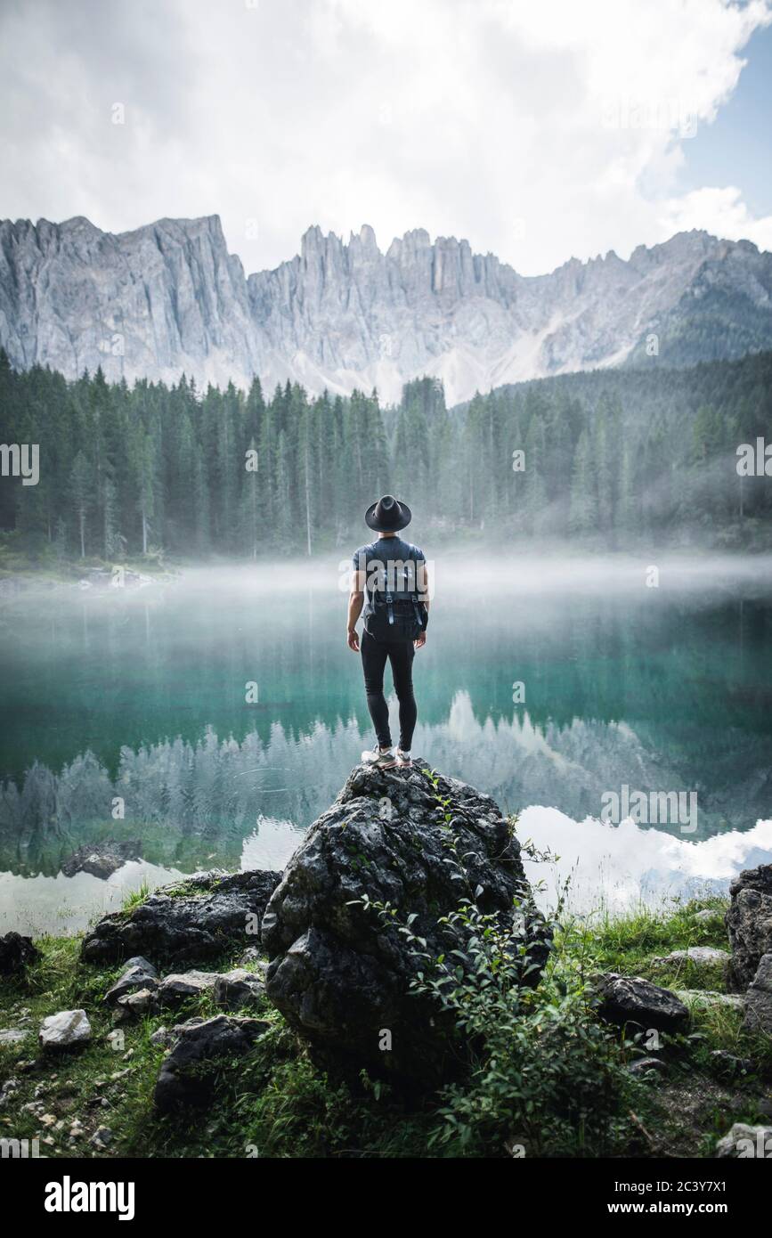 Italien, Karersee, junger Mann steht am Karersee in den Dolomiten bei Sonnenaufgang auf dem Felsen Stockfoto