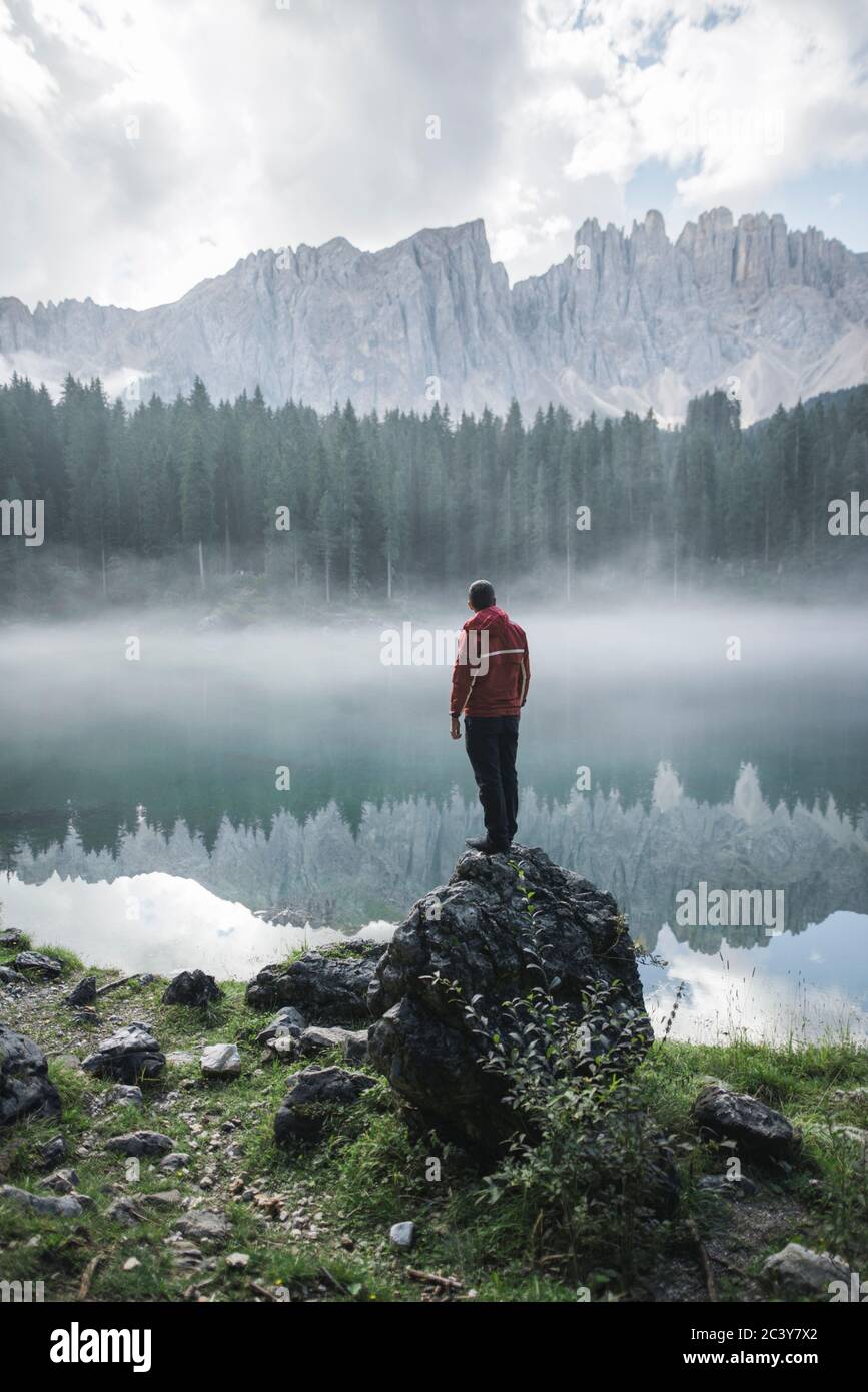 Italien, Karersee, junger Mann steht am Karersee in den Dolomiten bei Sonnenaufgang auf dem Felsen Stockfoto