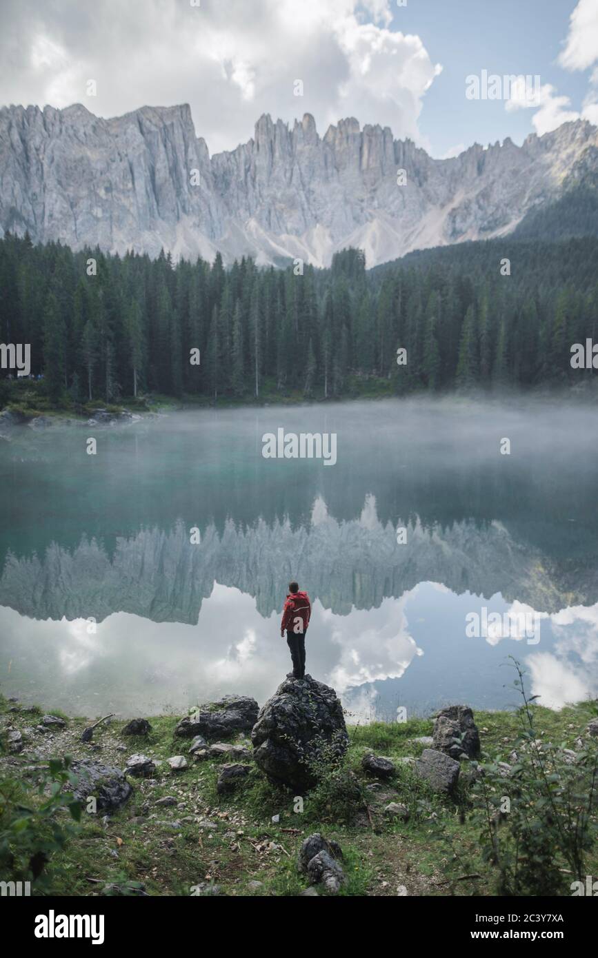 Italien, Karersee, junger Mann steht am Karersee in den Dolomiten bei Sonnenaufgang auf dem Felsen Stockfoto