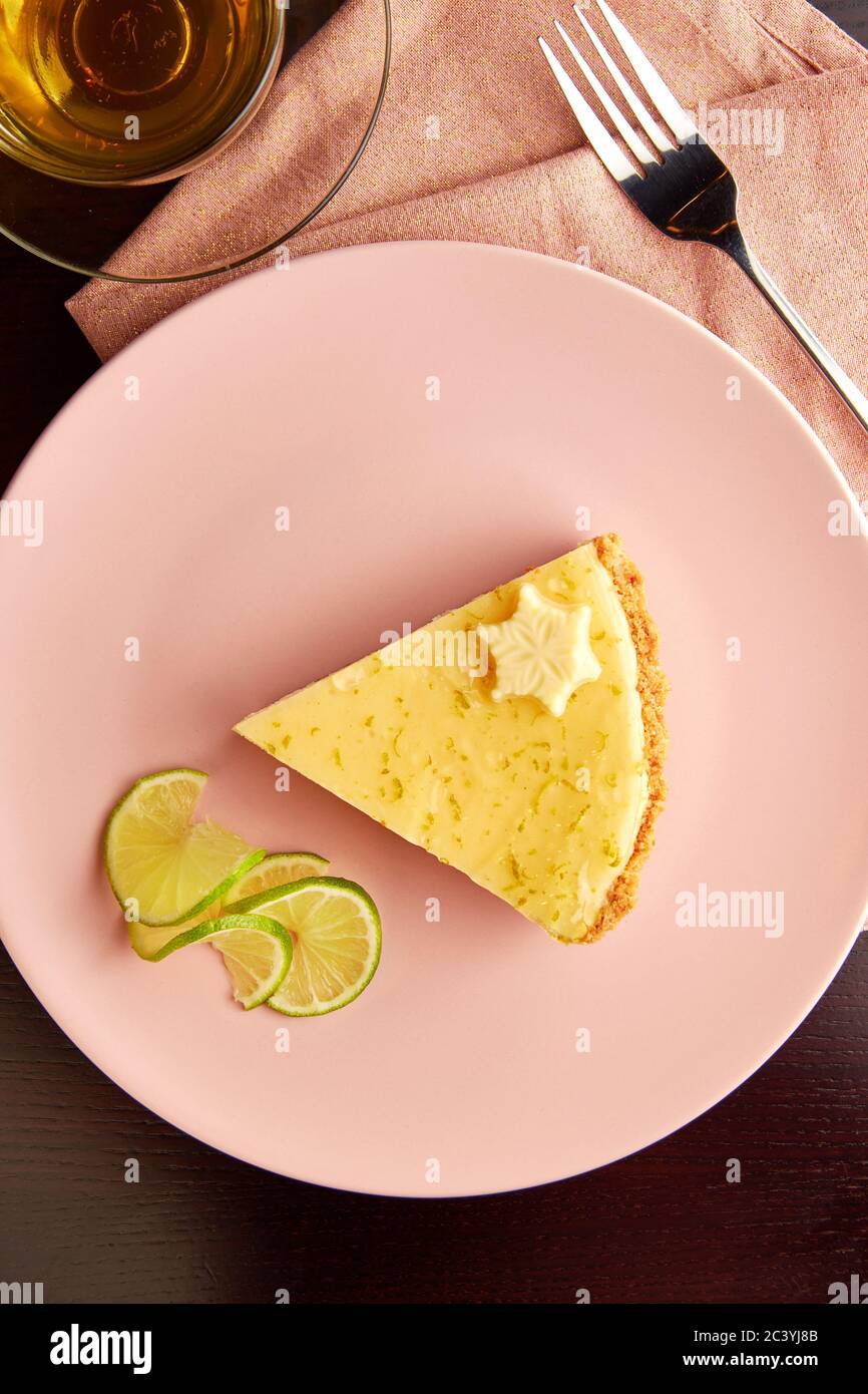 Stück leckeren Törtchen mit Limettenschale und Scheibe des Limes auf rosa Platte auf dunklen Holztisch, serviert mit Tasse Kräutertee. Stockfoto