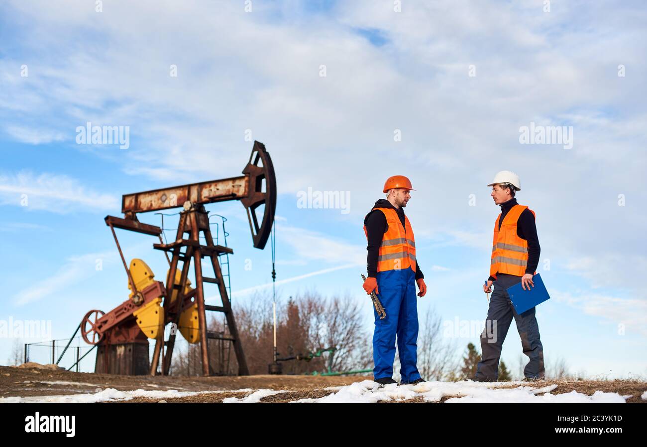 Zwei Ölmänner in Helmen und Arbeitswesten stehen in der Nähe von Ölbrunnen Pumpenheber und diskutieren Arbeit. Ölarbeiter hält Zwischenablage und spricht mit Kollegen auf dem Ölfeld. Konzept der Erdölindustrie. Stockfoto