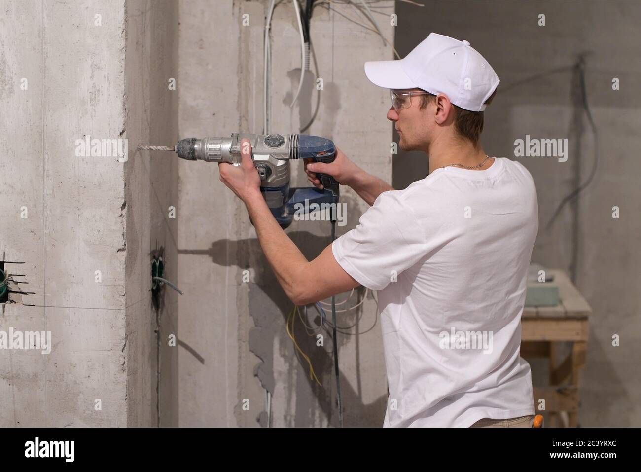Ein junger Arbeiter in weißer Uniform bohrt eine Wand Mit einem Schlag in der Wohnung Stockfoto