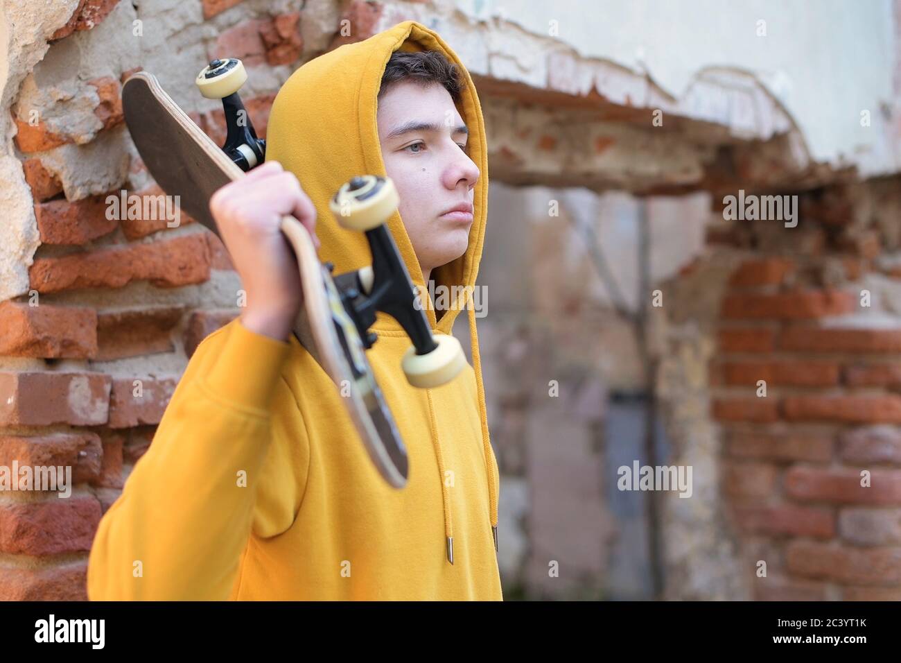 Ein Teenager mit einem Skateboard auf der Schulter steht an einer baufälligen Wand Stockfoto