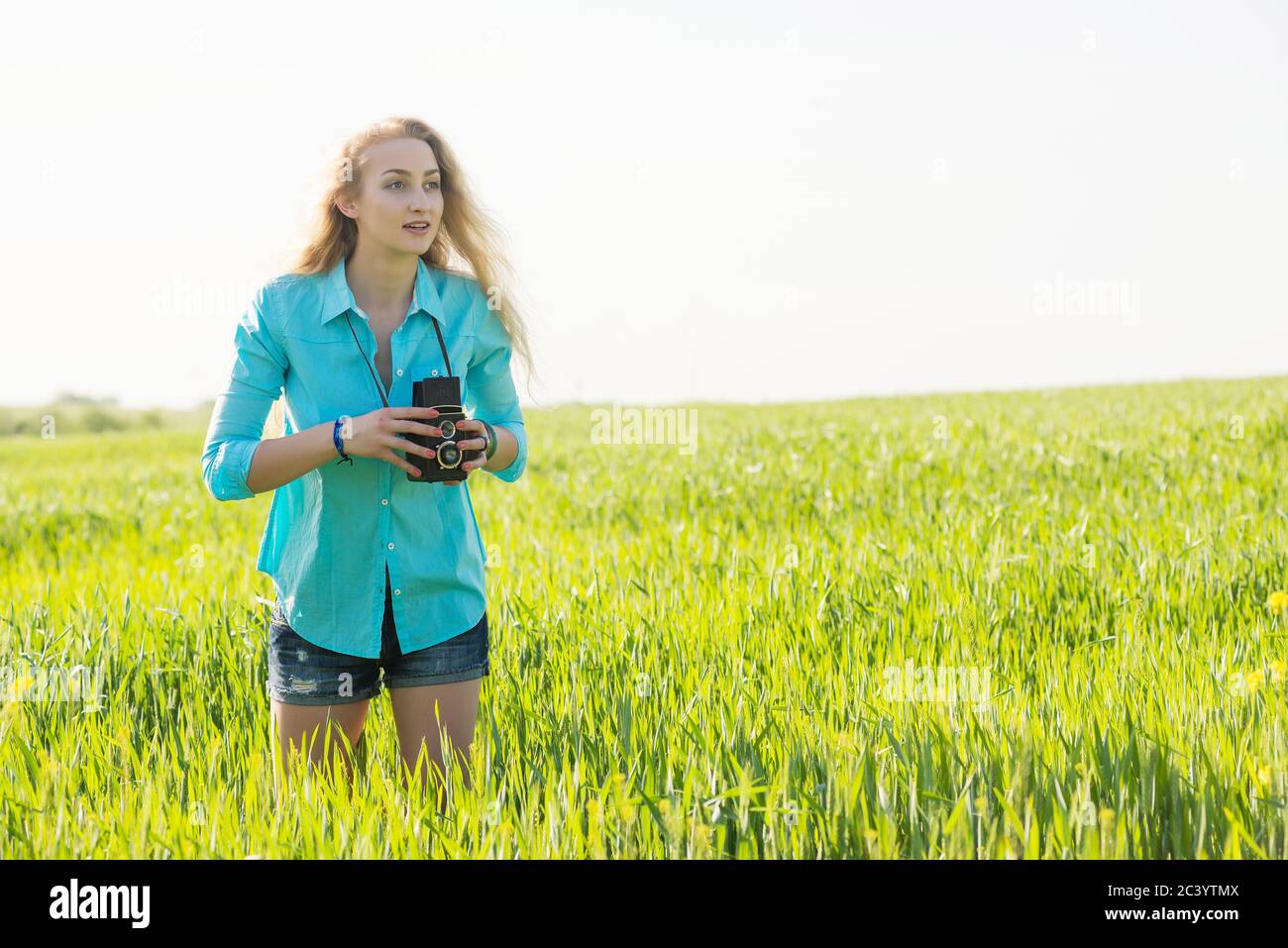 Junge blonde Frau mit Vintage-Kamera auf einem Sommer grünen Weizenfeld, die Inspiration fühlt und macht Bilder Stockfoto