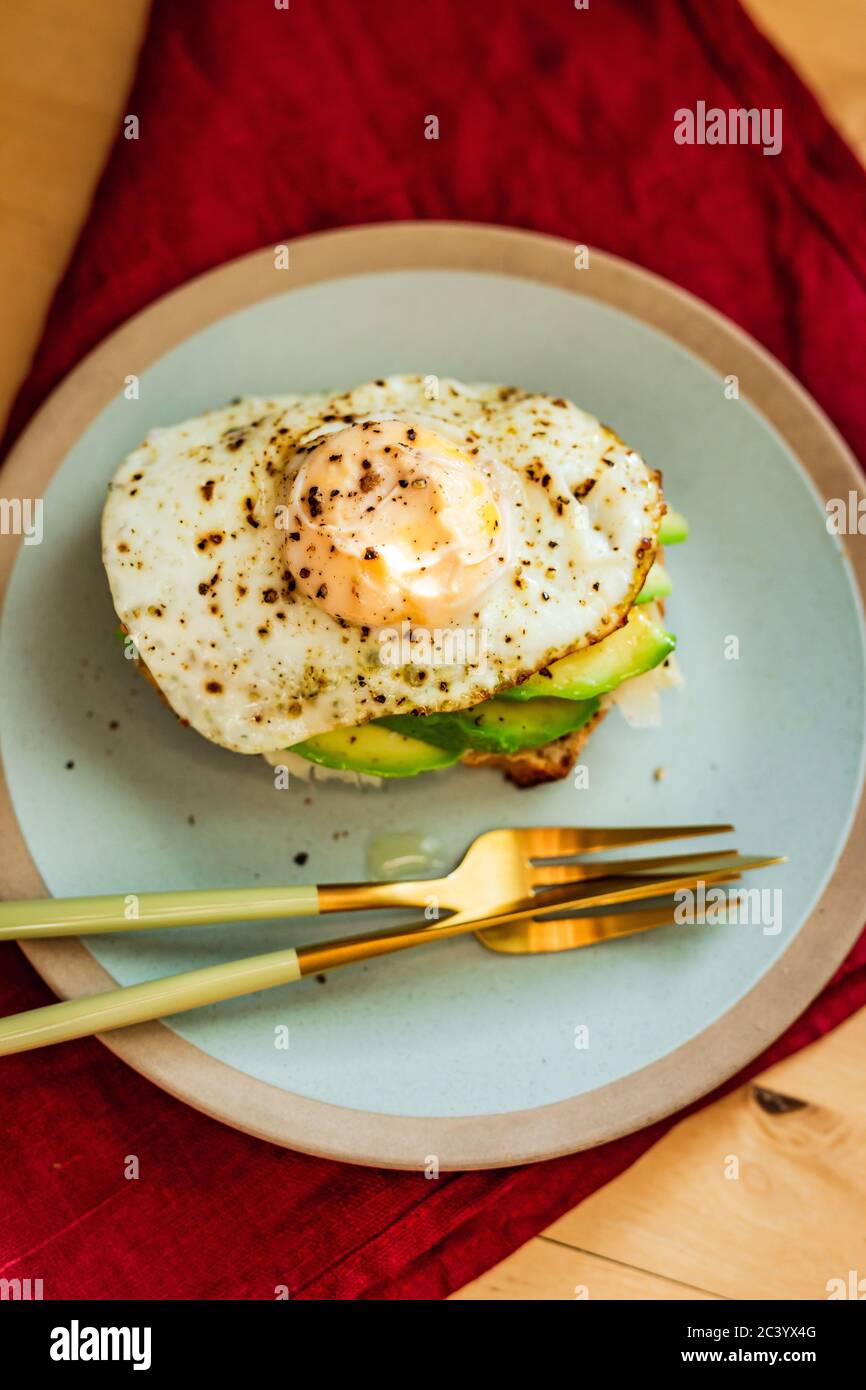 Ein Roggenbrot Toast mit Käse, Avocado und Spiegelei zum Frühstück, serviert auf einem blauen Teller. Schönes goldenes Besteck, rote Serviette. Stockfoto