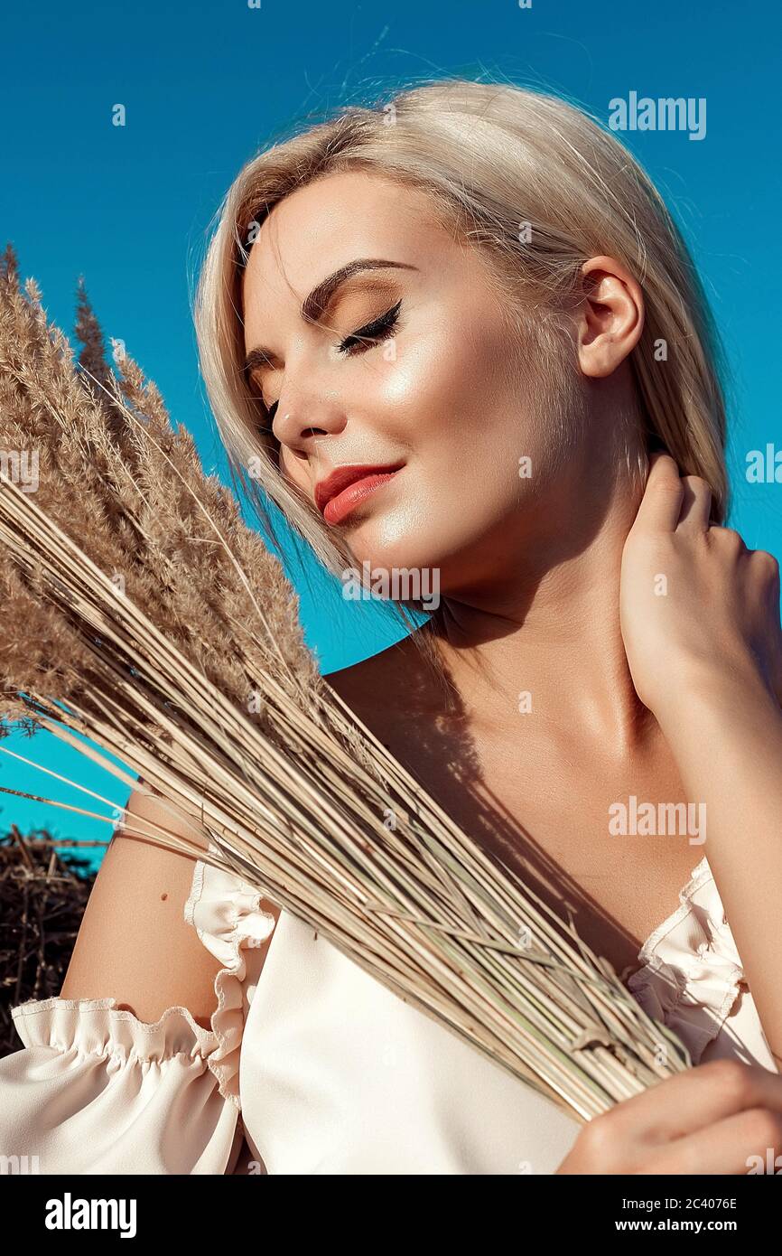 Portrait eines schönen blonden Mädchen mit langen Haaren auf einem Feld ein sonniger Tag bei Sonnenuntergang. In ihren Händen hält sie gelbes hohes und trockenes Gras. Auf Dem Land. Stockfoto