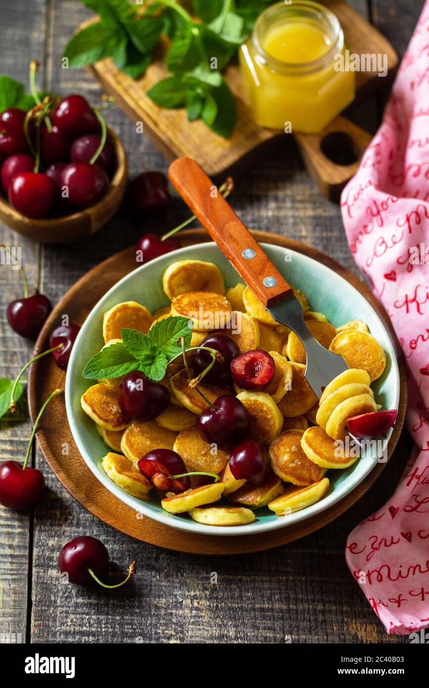 Winziges Pfannkuchen-Frühstück. Mini-Pfannkuchen mit Kirschen und Honig auf rustikalem Holztisch. Stockfoto