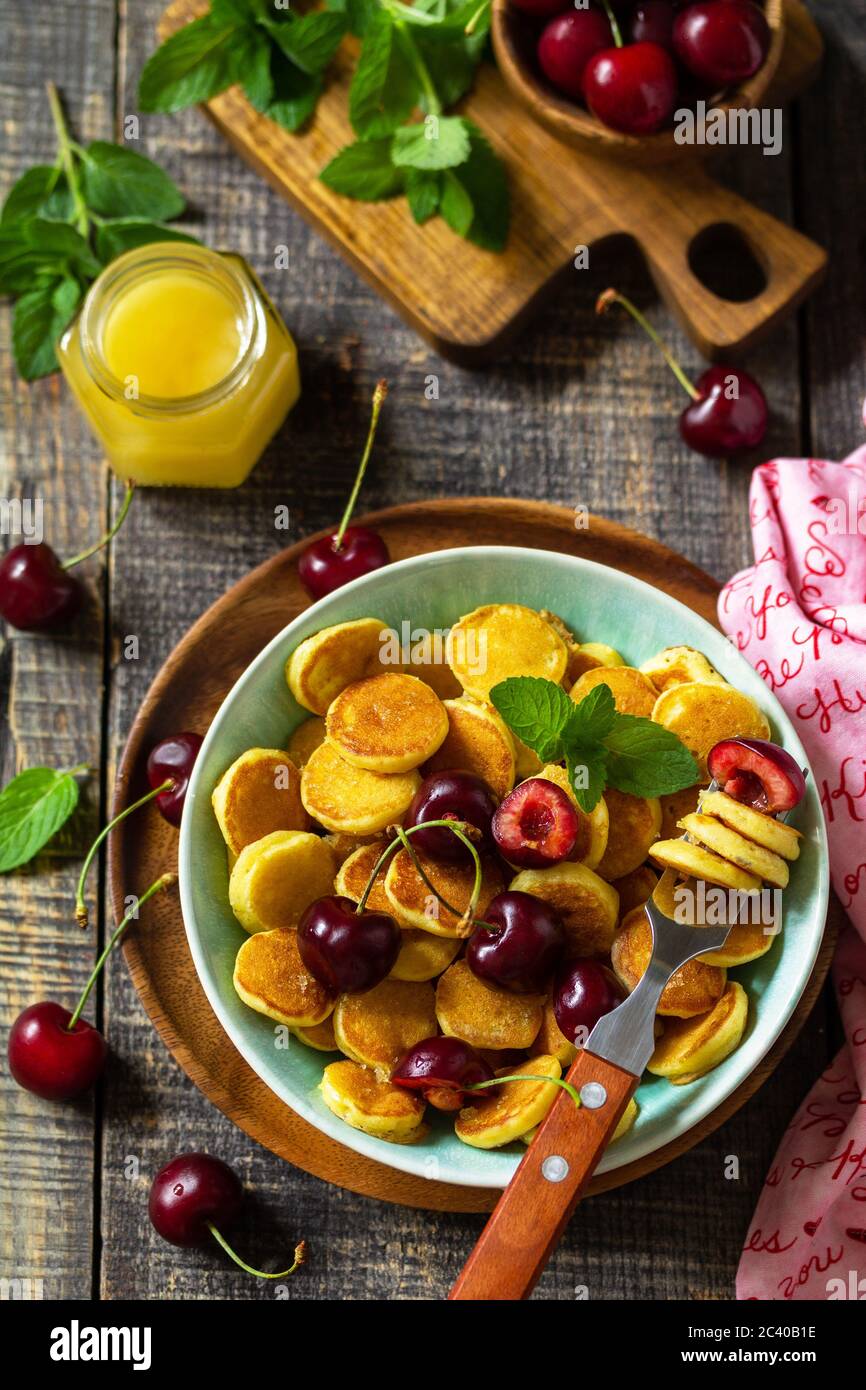 Winziges Pfannkuchen-Frühstück. Mini-Pfannkuchen mit Kirschen und Honig auf rustikalem Holztisch. Stockfoto