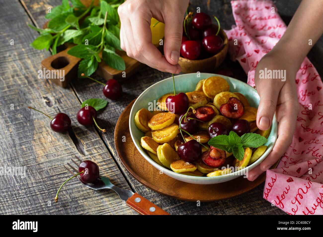 In den Händen einer Frau Frühstück. Mini-Pfannkuchen mit Kirschen und Honig auf einem rustikalen Holztisch. Stockfoto