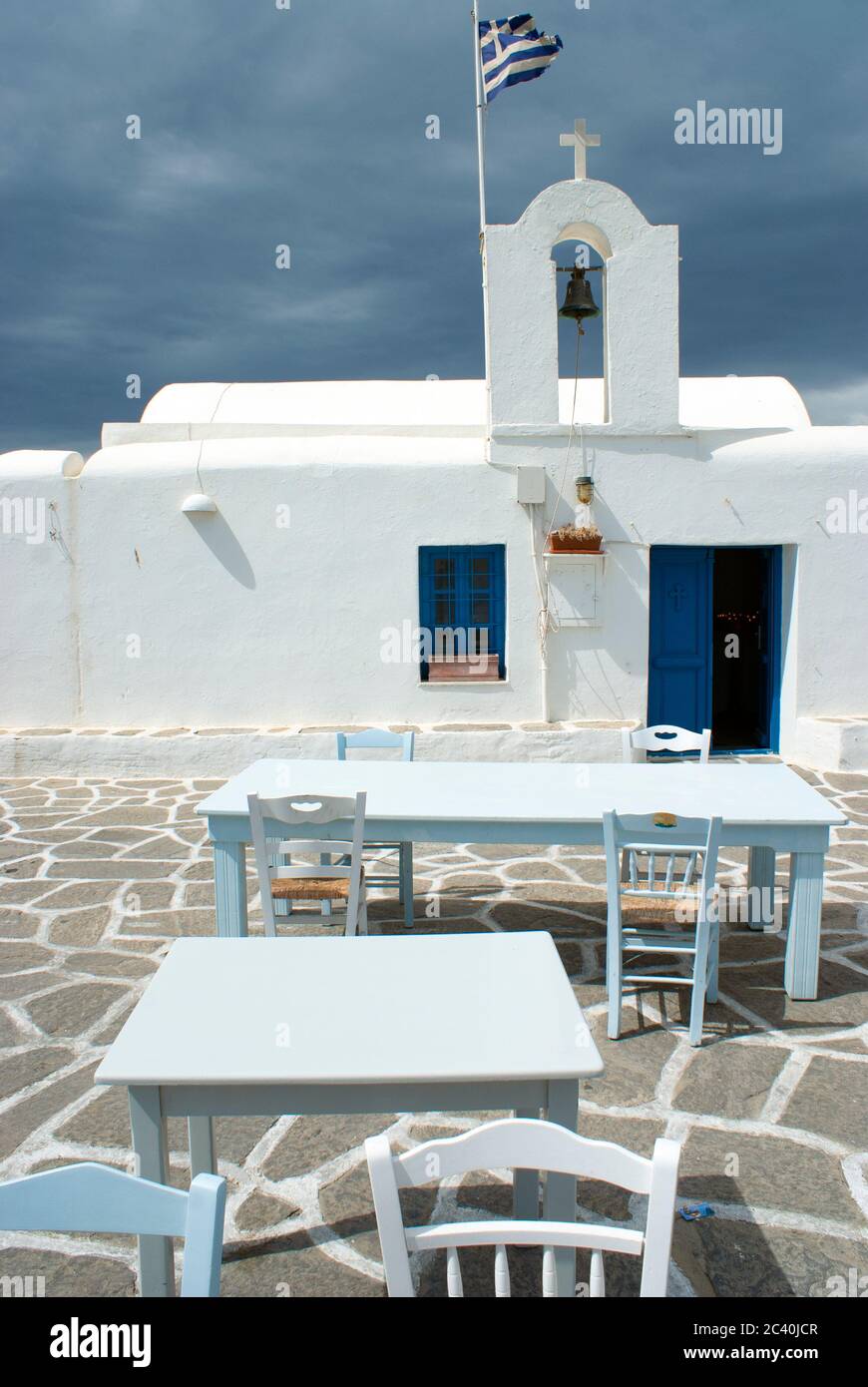 Auf der Insel Paros werden Tische und Stühle von einer Taverne auf der Terrasse durch eine altmodische orthodoxe Kapelle aufgestellt. Sturmwolken sammeln sich. Stockfoto