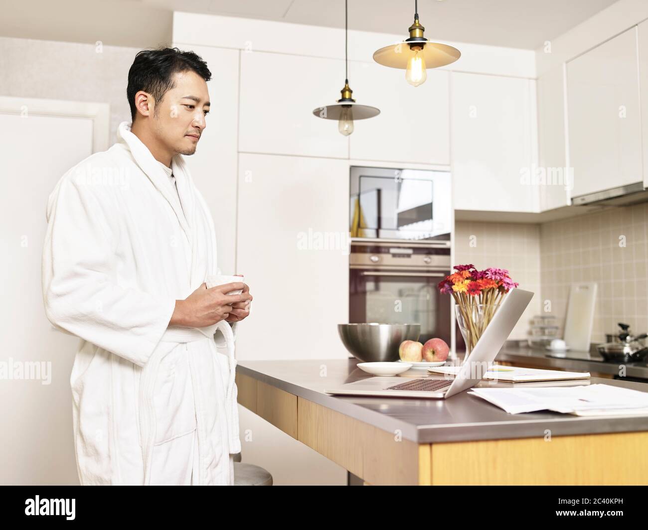 Junger asiatischer Geschäftsmann im Bademantel, der von zu Hause aus an der Küchentheke steht und eine Tasse Kaffee hält und dabei den Laptop anschaut Stockfoto