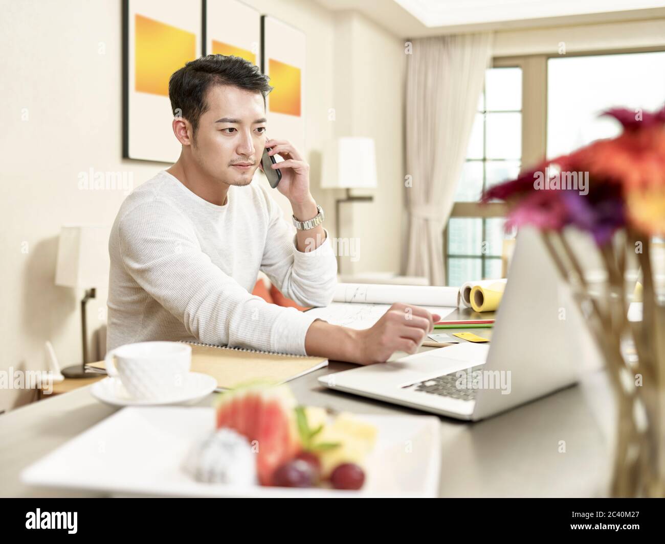Junge asiatische Mann Design professionelle Arbeit von zu Hause sitzen in der Küche sprechen auf dem Handy, während Blick auf Laptop-Computer (Kunstwerk im Hintergrund Stockfoto