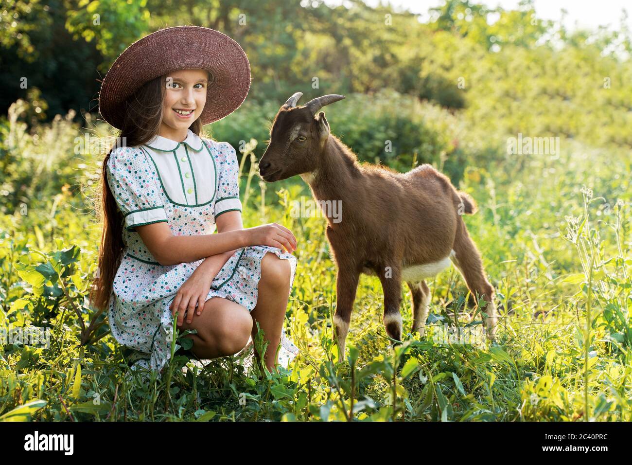 Ein kleines Mädchen in einem Hut hütet eine Ziege In einer grünen Wiese Stockfoto