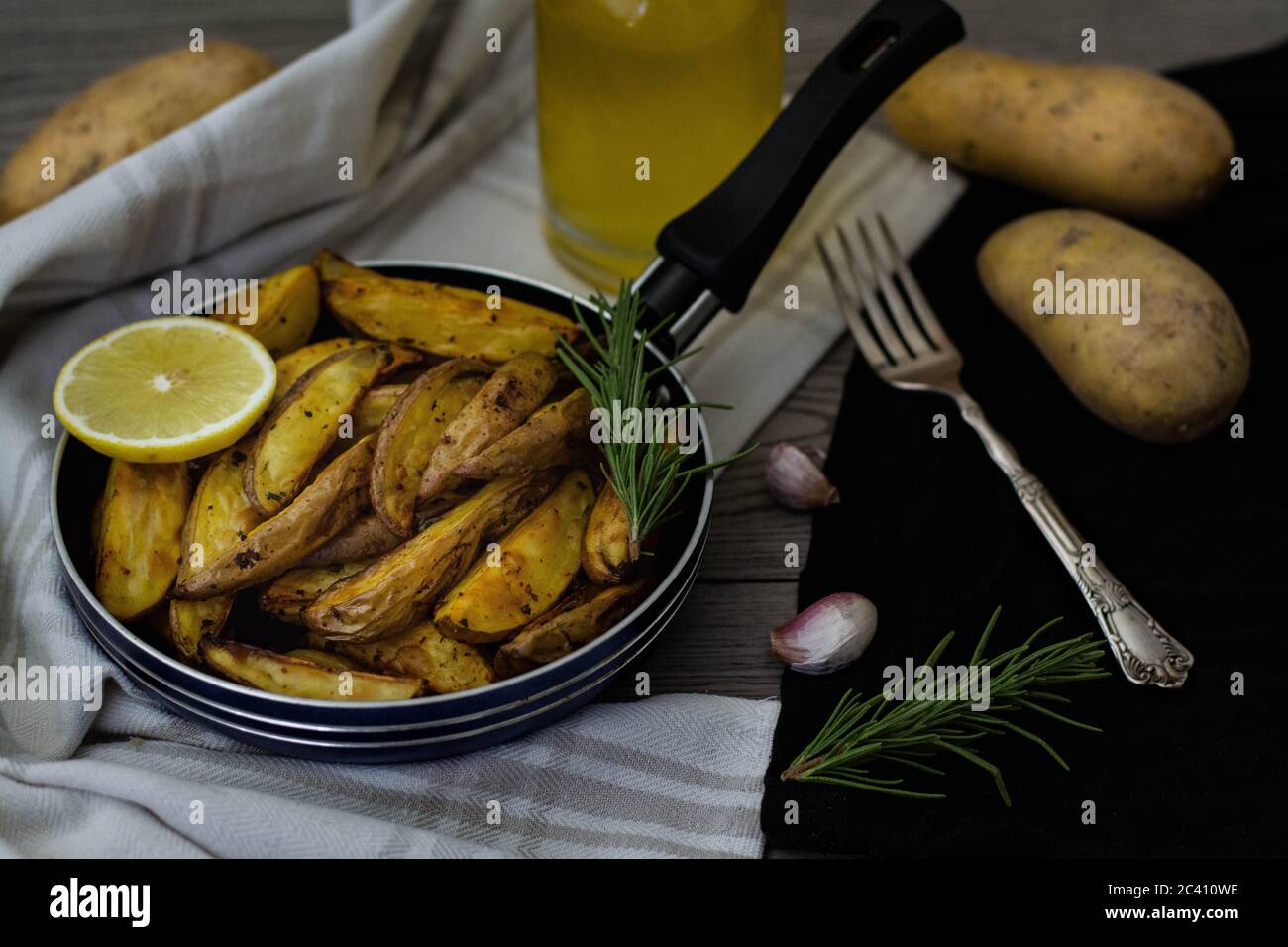 Gebackene Kartoffelkeile mit Rosmarin, Knoblauch und Zitrone in einer kleinen Pfanne. Rohe Kartoffeln, Vintage Silbergabel, Rosmarin und Olivenöl im Hintergrund. Stockfoto