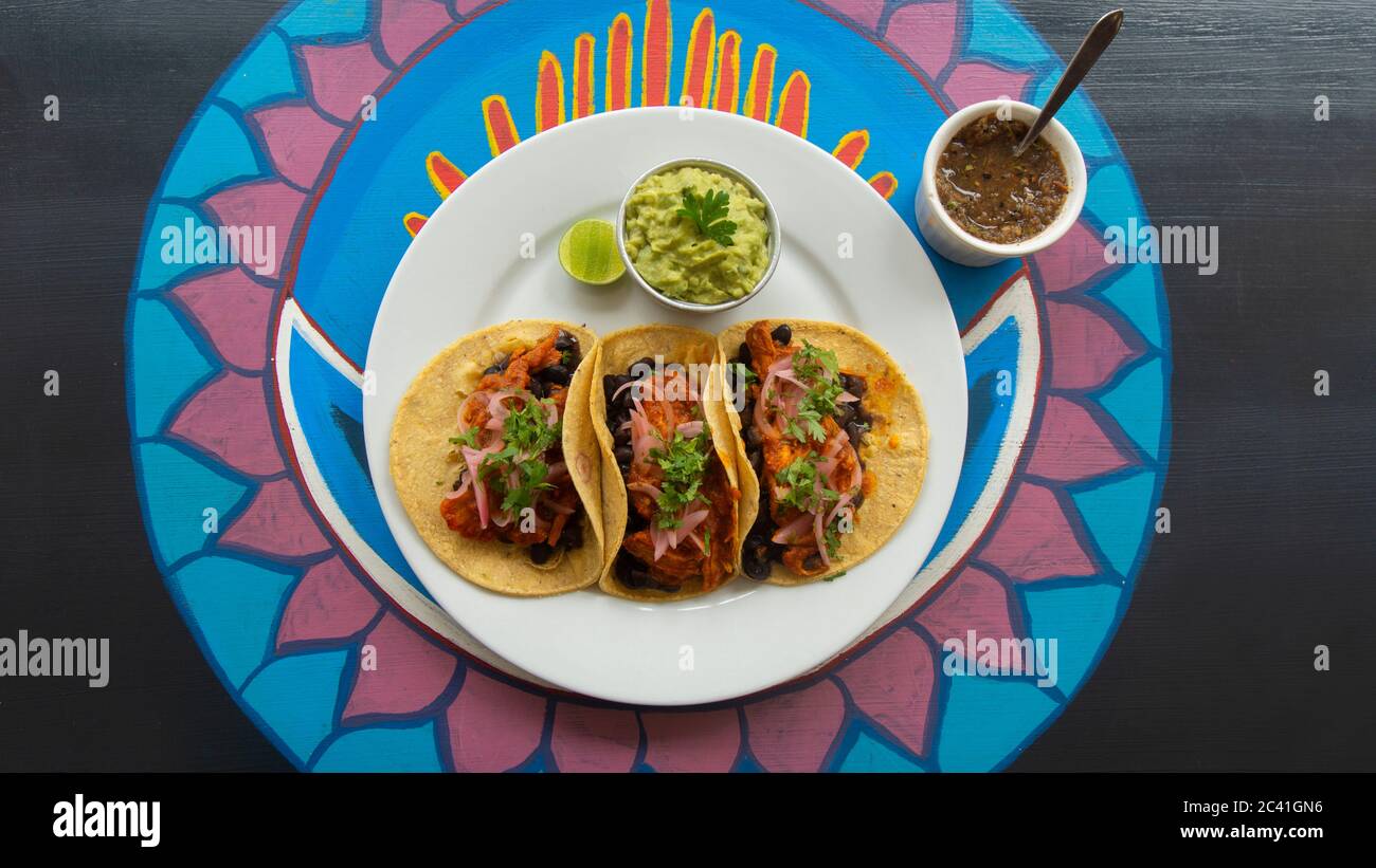 Draufsicht auf einen weißen Teller mit drei mexikanischen Tacos Fleisch, begleitet von einem kleinen Topf Guacamole und würziger Sauce auf einem Holztisch mit verschiedenen Colos Stockfoto