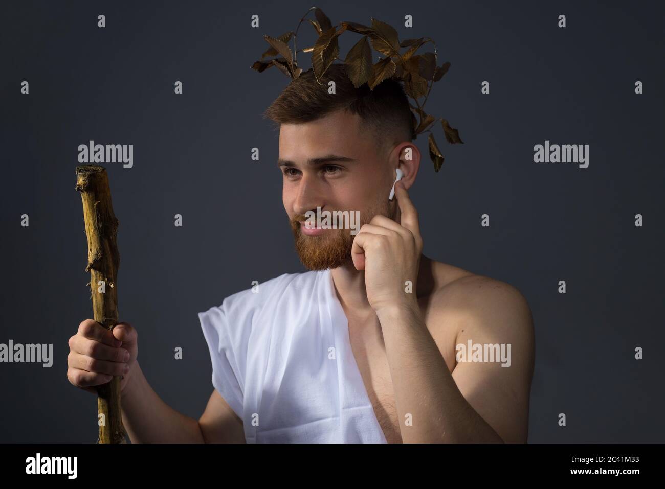 Altgriechischer bärtiger gott in kabellosen Kopfhörern, hört Musik, in einem Lorbeerkranz mit einem Stab. Studio Foto in dramatischem Licht, auf einem grauen Stockfoto