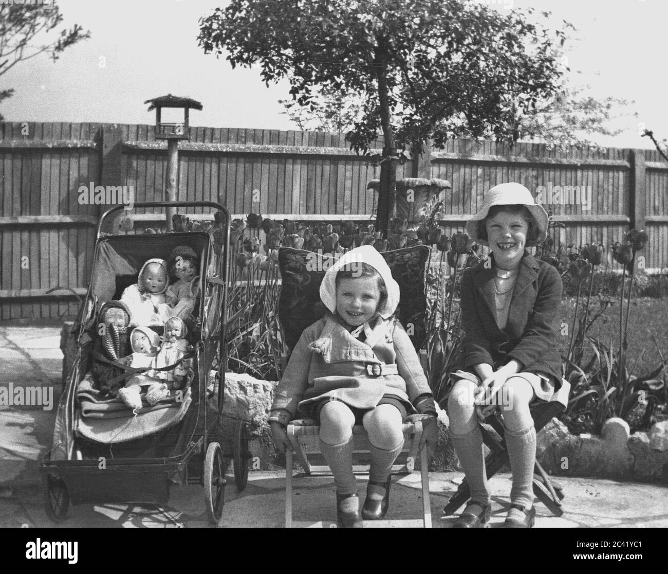 1930er Jahre, historiacl, zwei glückliche junge Mädchen, möglicherweise Schwestern, sitzen draußen in einem Garten neben einem kleinen Metall gerahmten Kinderwagen mit ihren Spielzeugpuppen, England, Großbritannien. Stockfoto