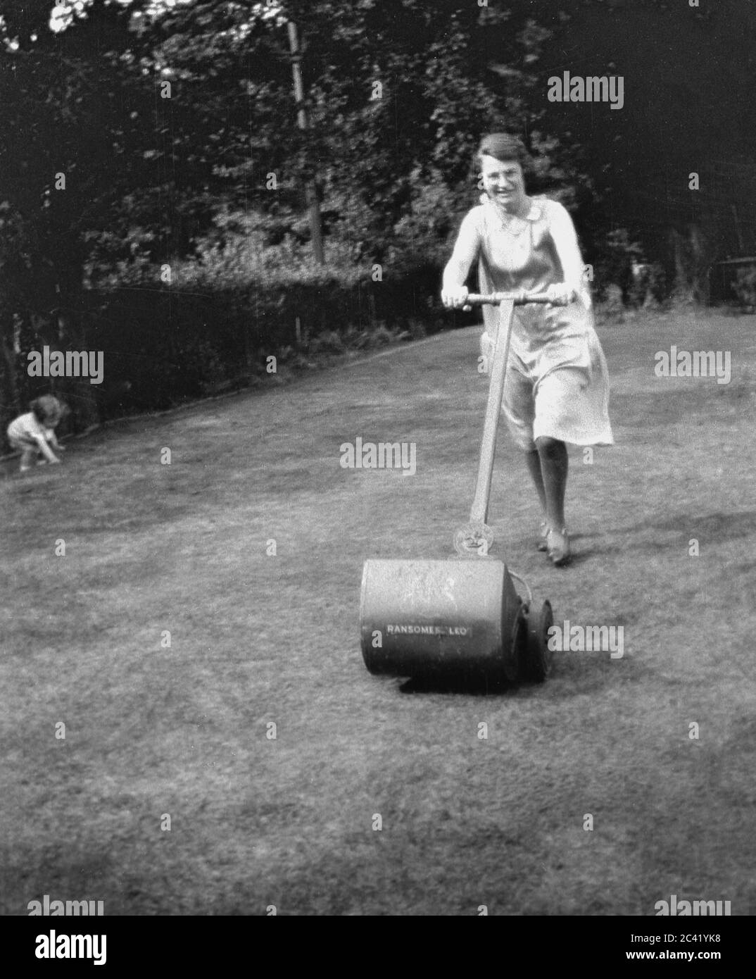 1930er Jahre, historisch, eine Dame, die in einem Garten mit Rasen-Mover der Zeit das Gras schneidet, mit ihrem Kleinkind, das in der Ecke spielt, England, Großbritannien. Sie verwendet einen Handmäher von Ransomes, der 1832 den ersten Rasenmäher der Welt hergestellt hat, unter Lizenz einer Erfindung, die 1830 von Edward Beard einem Ingenieur aus Stroud, Gloucestershire, England, geboren wurde. Stockfoto