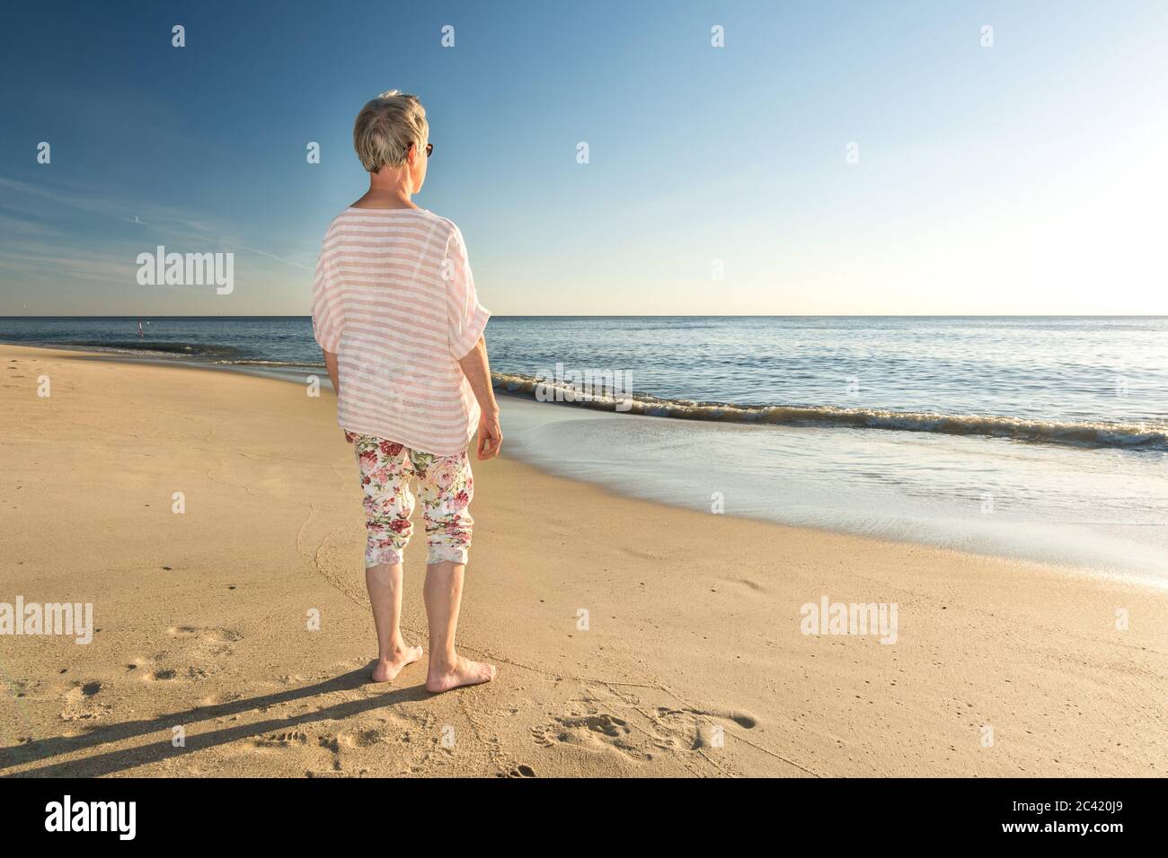Ältere Frau, die am Strand steht und auf das Meer blickt Stockfoto