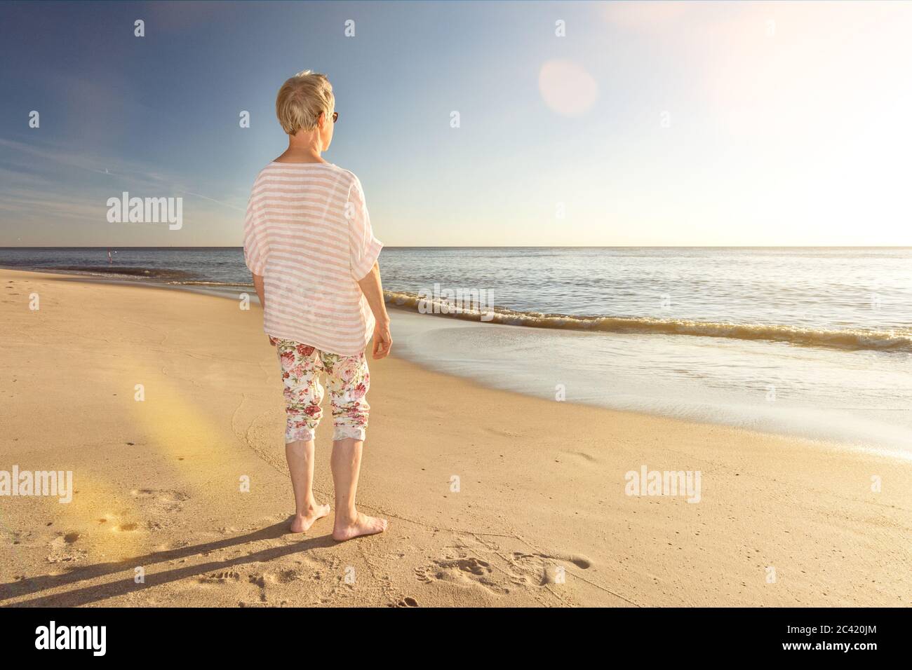 Ältere Frau, die am Strand steht und mit einer malerischen Linse auf das Meer blickt Stockfoto