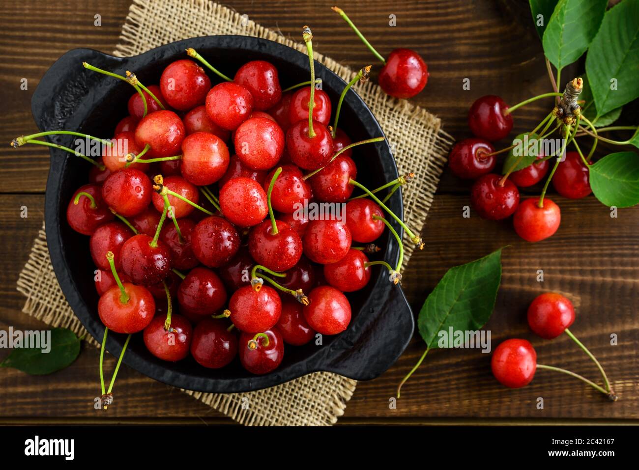 Frische süße rote Kirschen in einer schwarzen Schale und grüne Blätter auf einem dunkelbraunen Holztisch. Draufsicht. Stockfoto