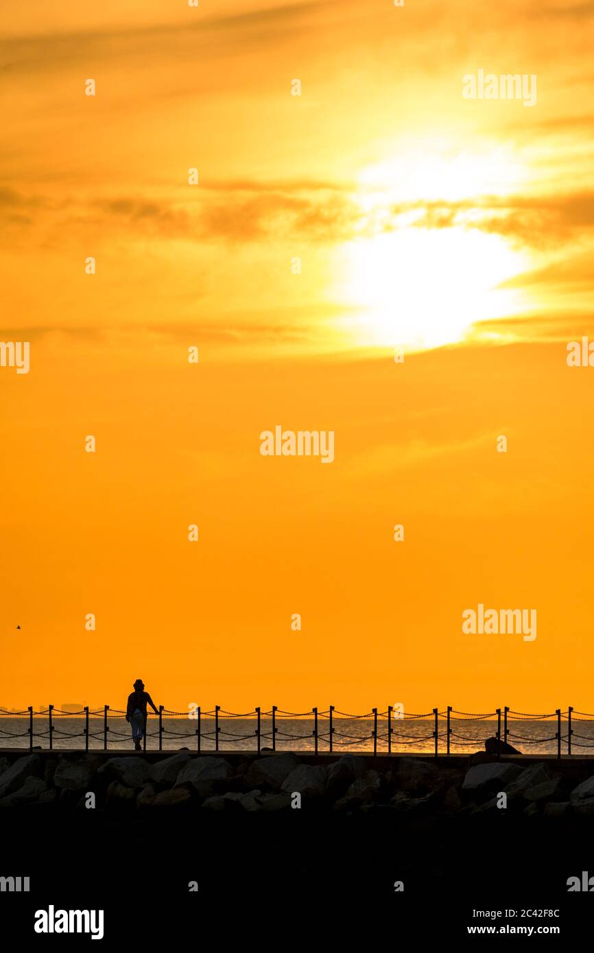Frau mittleren Alters hat sich gegen den Sonnenuntergang orange Himmel zu Fuß allein entlang Hafenmauer mit Kettengeländer in Herne Bay in Kent. Sonne am Seil des Rahmens, Frau und Geländer unten. Wolkiger Sonnenuntergang oranger Himmel. Stockfoto