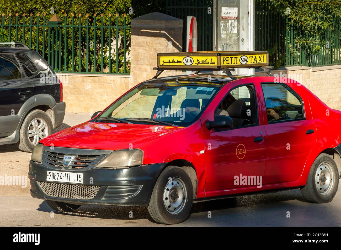 Ein rotes Petit-Taxi mit einem gelben Dachschild mit der Aufschrift „504“ parkt vor einem grünen Zaun in Fès, Marokko, neben ihm steht ein schwarzer SUV. Stockfoto