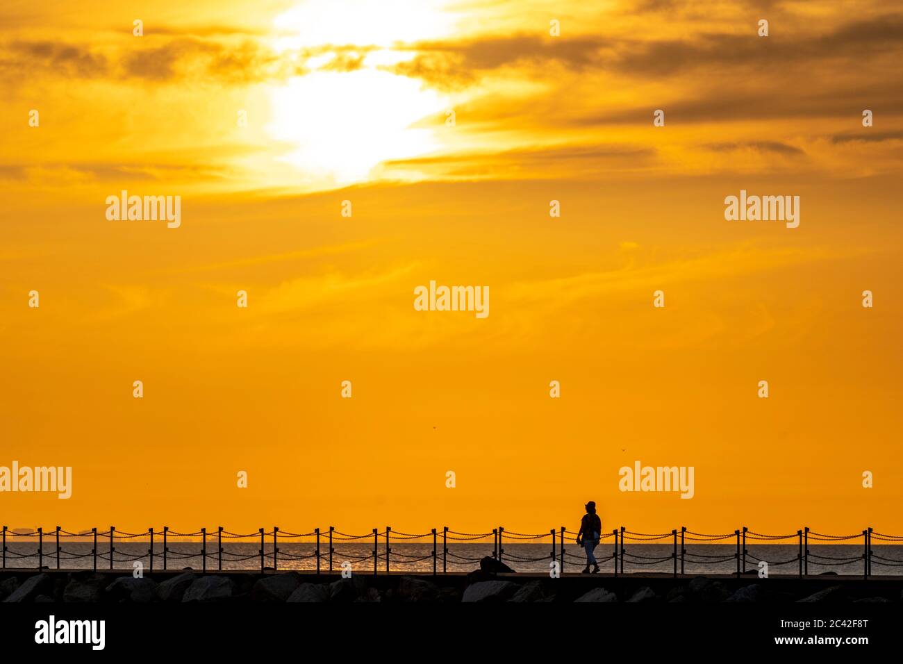 Frau mittleren Alters hat sich gegen den Sonnenuntergang orange Himmel zu Fuß allein entlang Hafenmauer mit Kettengeländer in Herne Bay in Kent. Sonne am Seil des Rahmens, Frau und Geländer unten. Wolkiger Sonnenuntergang oranger Himmel. Stockfoto