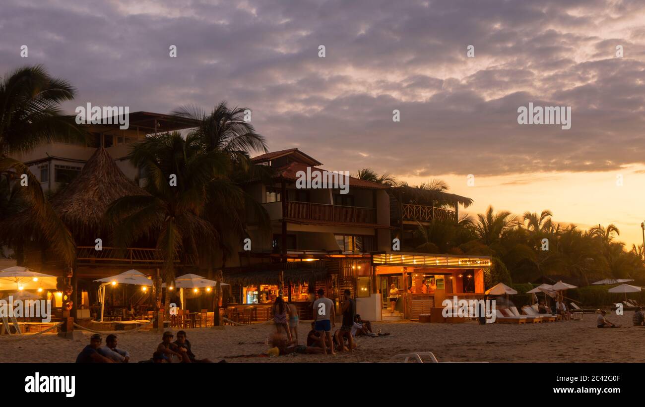 Mancora, Piura / Peru - 7. April 2019: Touristen, die am Strand spazieren gehen, mit den Lichtern des Dorfes bei Sonnenuntergang beleuchtet Stockfoto