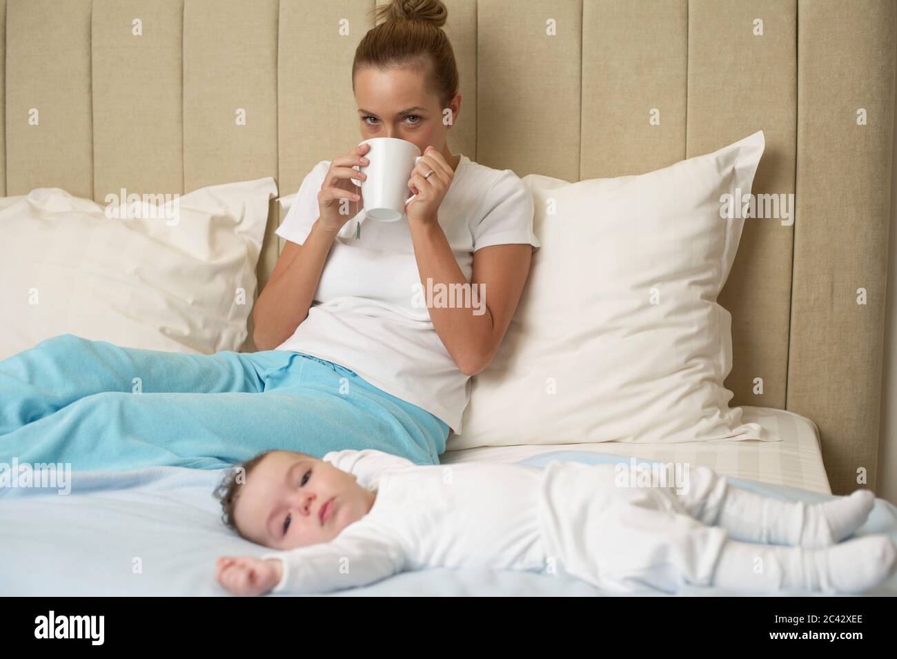 Mutter sitzt auf dem Bett und trinkt eine Tasse Tee, Baby steht im Vordergrund Stockfoto