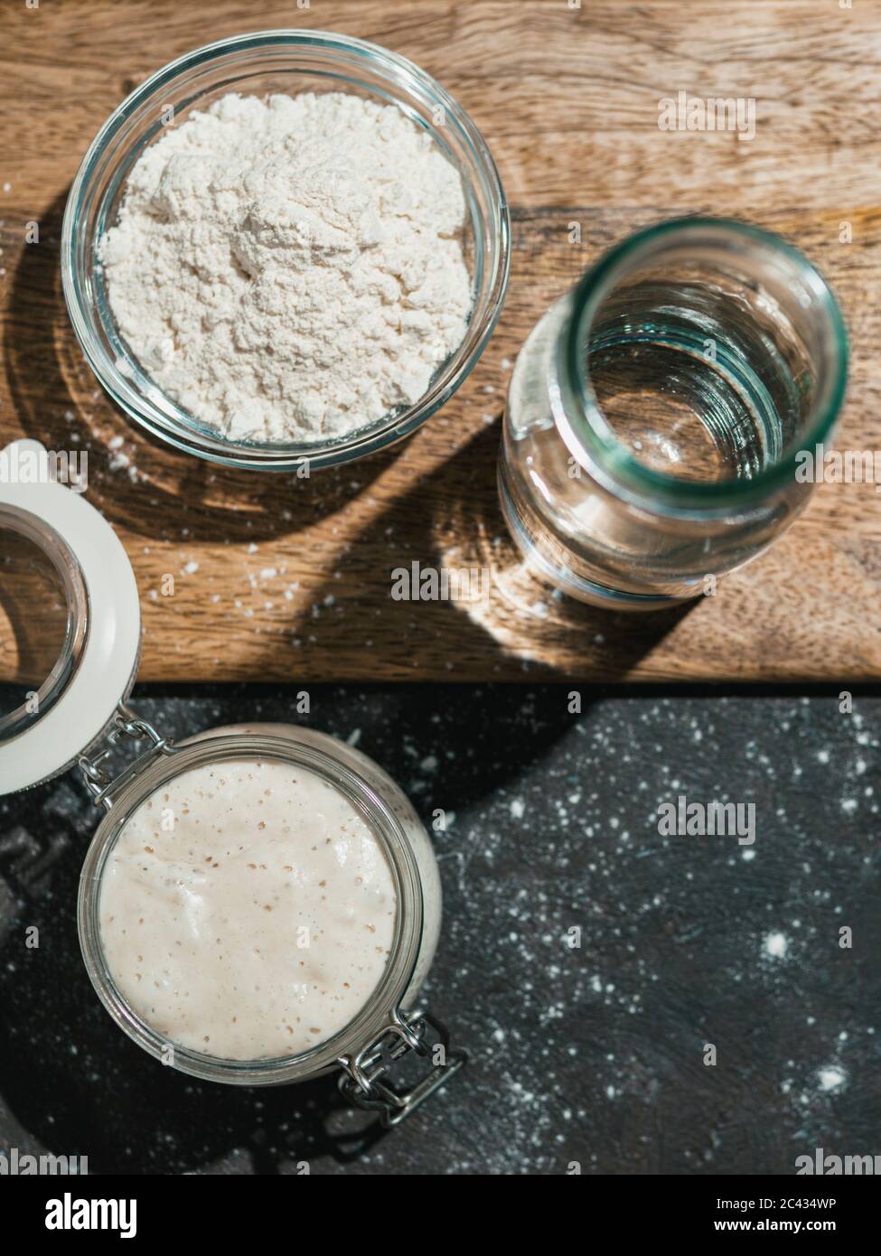 Weizensauerteigstarter in hartem Licht. Ansicht von oben auf Glas mit Sauerteig Starter, Wasserflasche und Weizenmehl Stockfoto