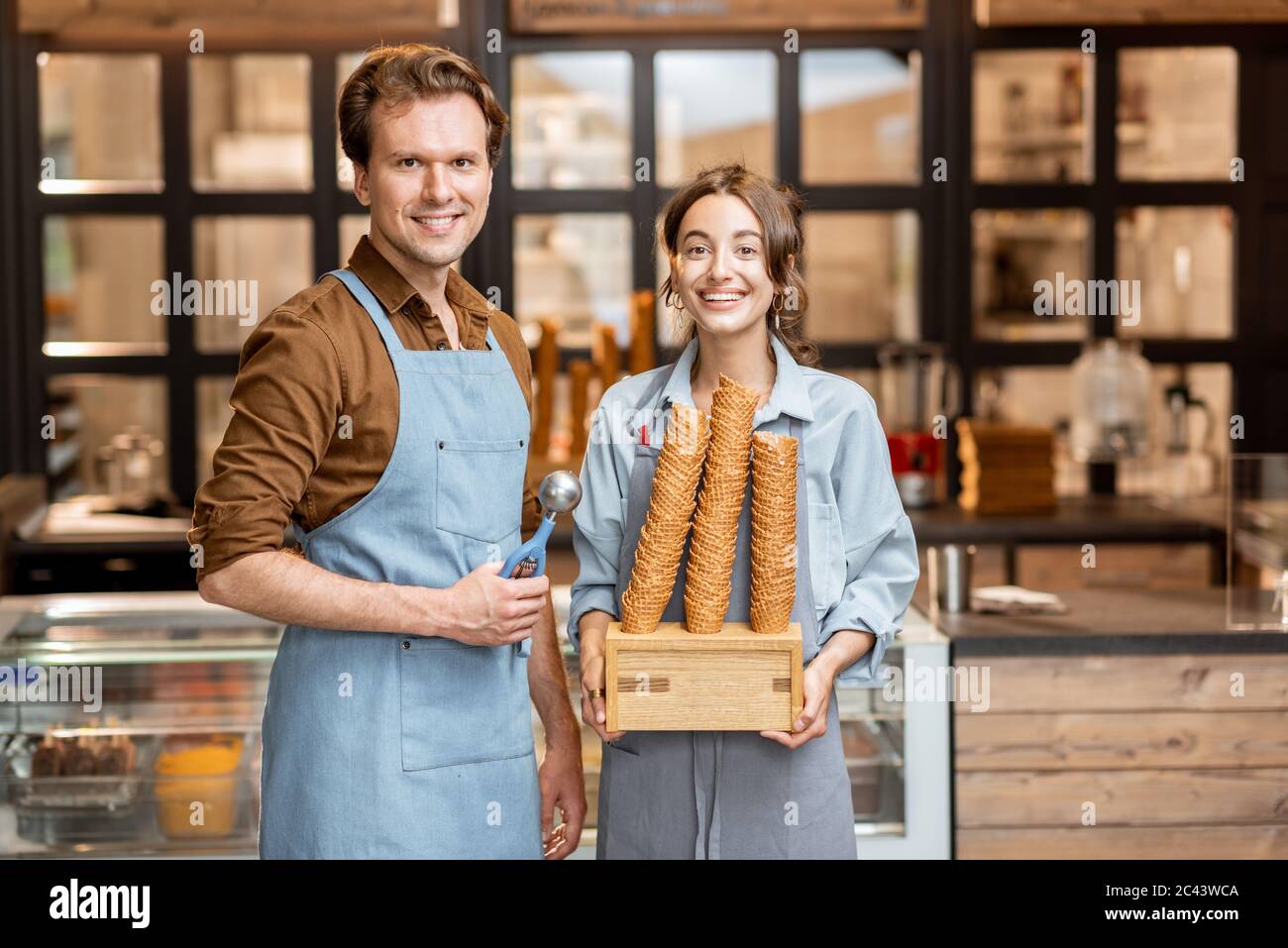 Porträt von zwei fröhlichen Mitarbeitern eines Cafés oder einer kleinen Konditorei, die mit einem Eiszapfen vor der Theke im Inneren stehen Stockfoto