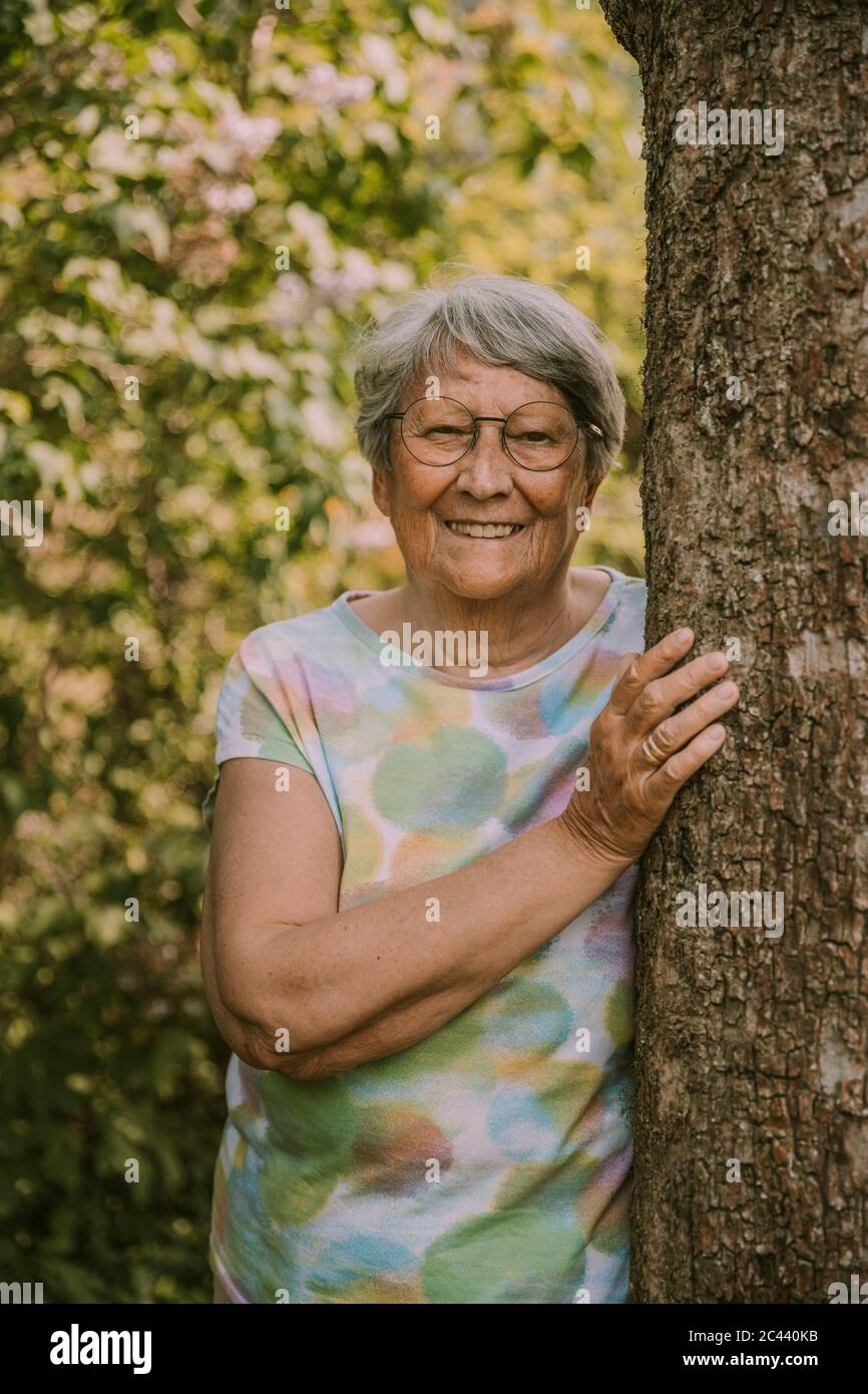 Glückliche ältere Frau im Ruhestand, die am Baum gegen Pflanzen im Garten steht Stockfoto