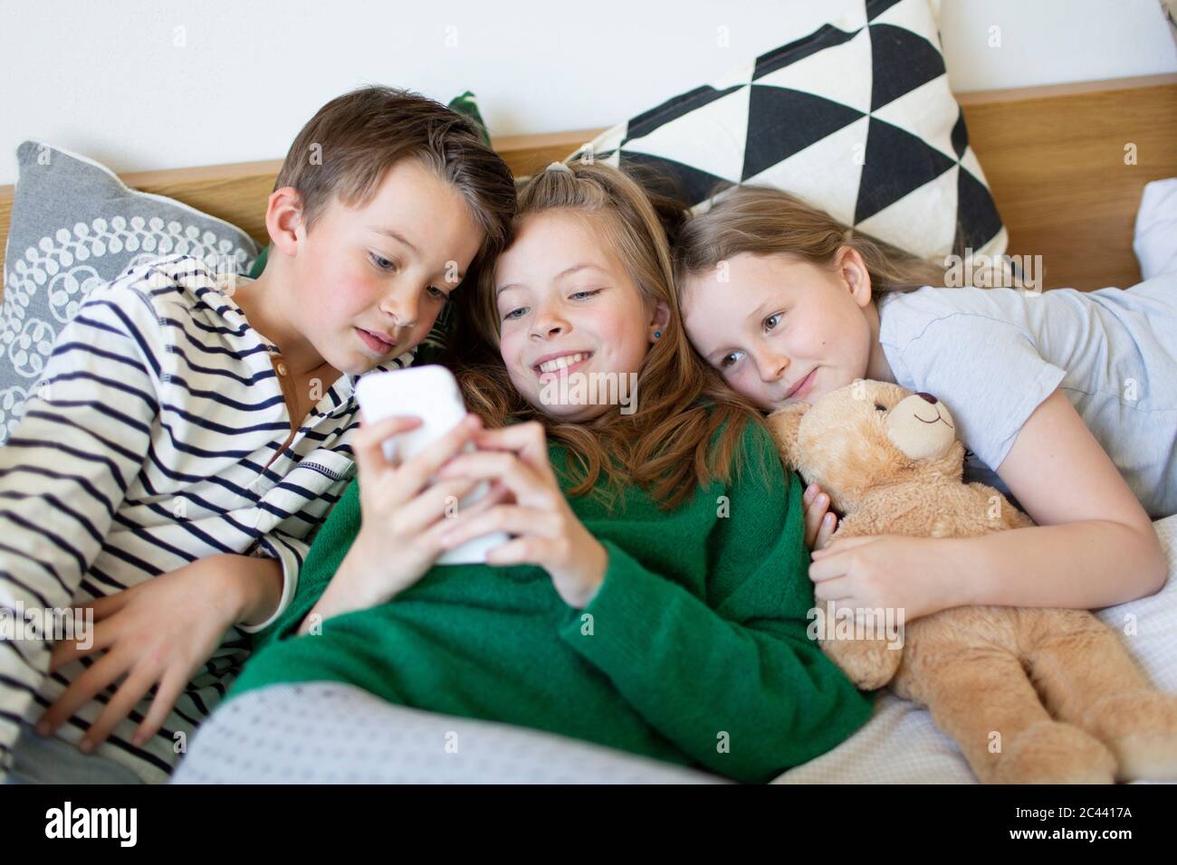 Gruppenbild von drei Kindern, die zusammen auf dem Bett liegen und auf das Handy blicken Stockfoto