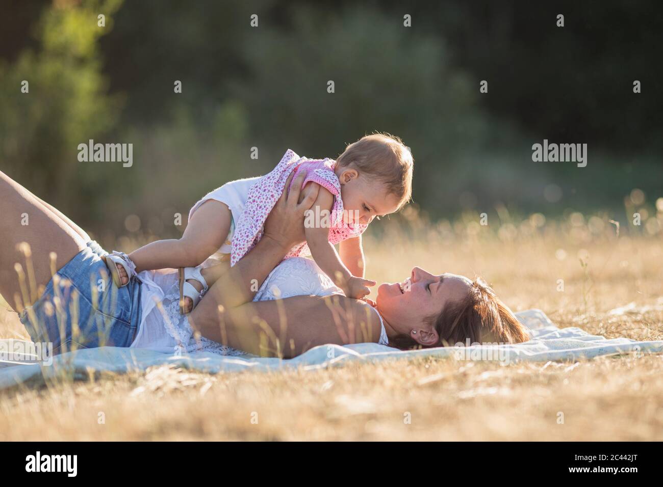 Glückliche Frau trägt Baby Mädchen, während auf Decke auf der Wiese während sonnigen Tag liegen Stockfoto