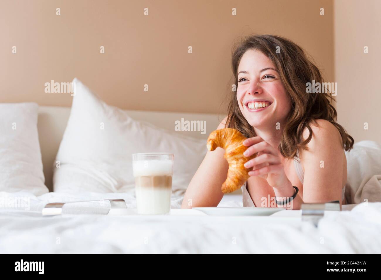 Glückliche junge Frau, die Croissant hält, während sie im Bett frühstückt Stockfoto