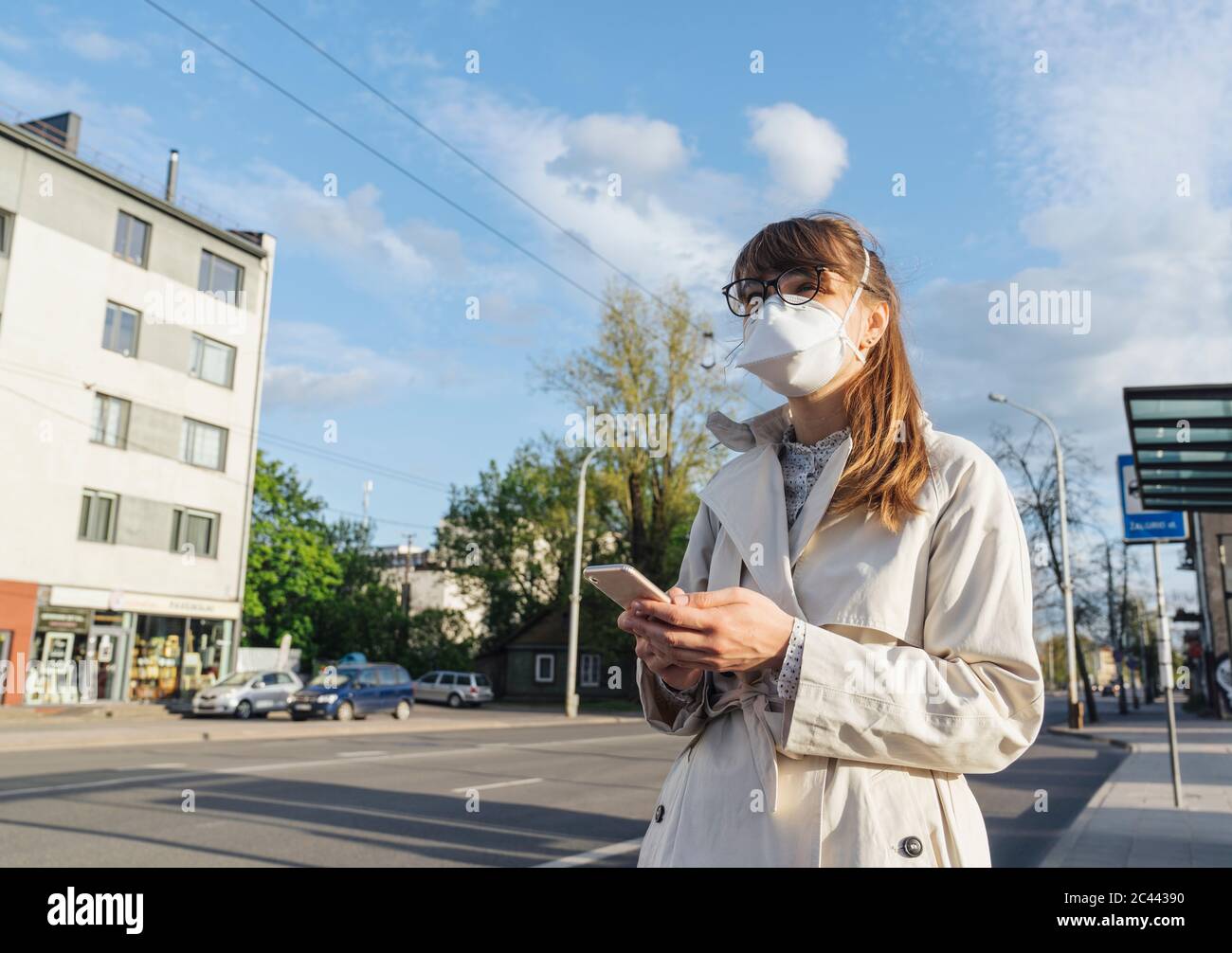 Frau trägt Gesichtsmaske und hält Smartphone wartet auf einen öffentlichen Verkehr in der Stadt Stockfoto