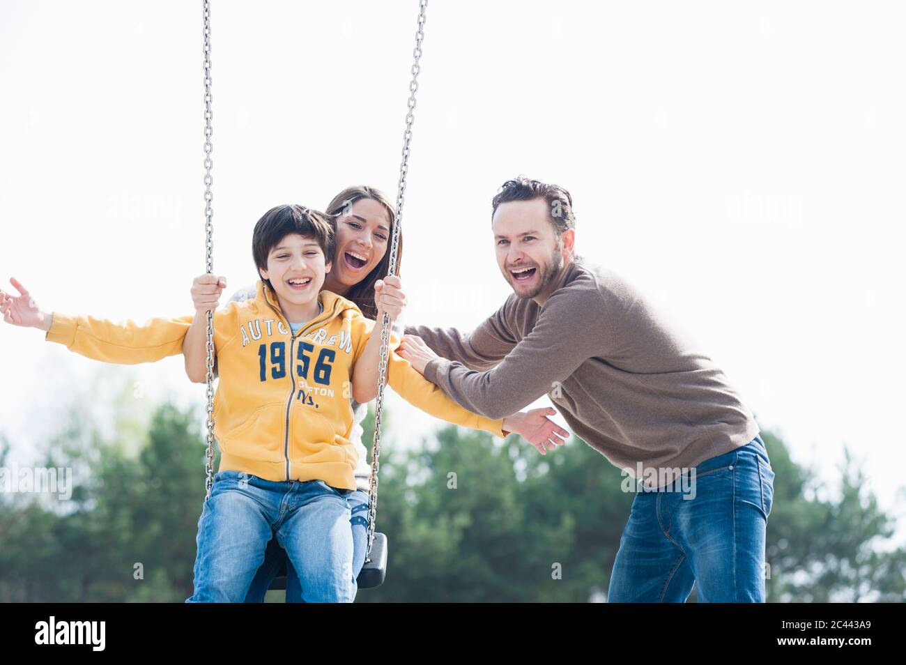 Fröhlicher Mann, der Mutter und Sohn auf Schaukel im Park gegen den klaren Himmel schiebt Stockfoto