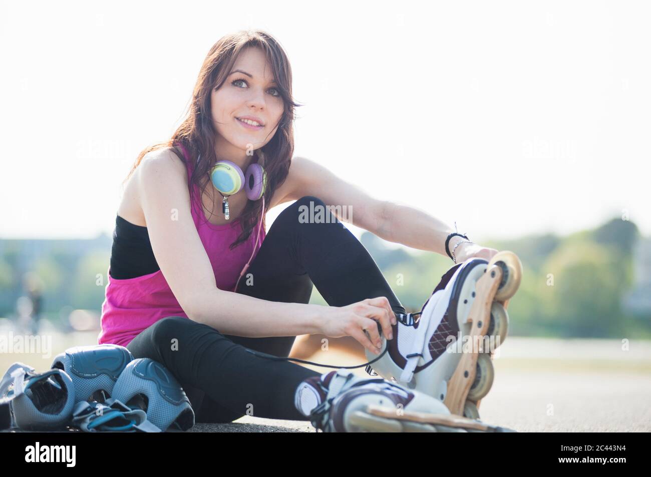Lächelnde, selbstbewusste junge Frau, die Inline-Skates trägt, während sie im Park gegen den klaren Himmel sitzt Stockfoto