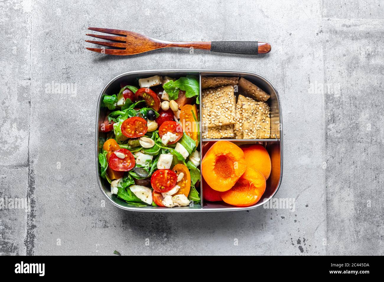 Lunchbox mit Rucola-Salat mit farbigen Tomaten, Mozzarella und Nüssen, Knäckebrot und Aprikosen, Holzgabel auf Betonoberfläche Stockfoto