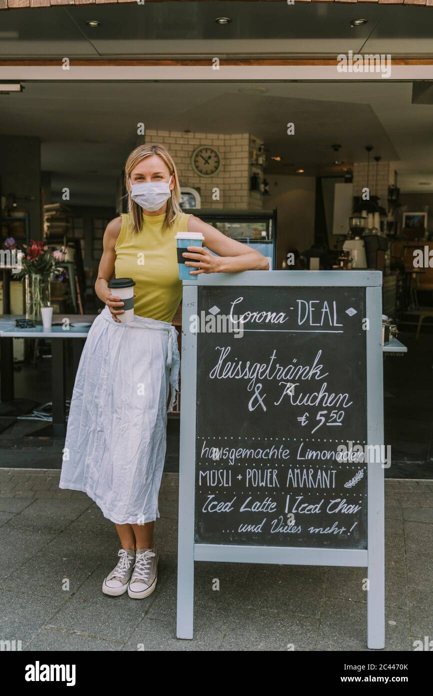 Frau trägt Gesichtsmaske und hält Kaffee zu gehen, während an einem Schild mit Corona Sonderangebote gelehnt Stockfoto