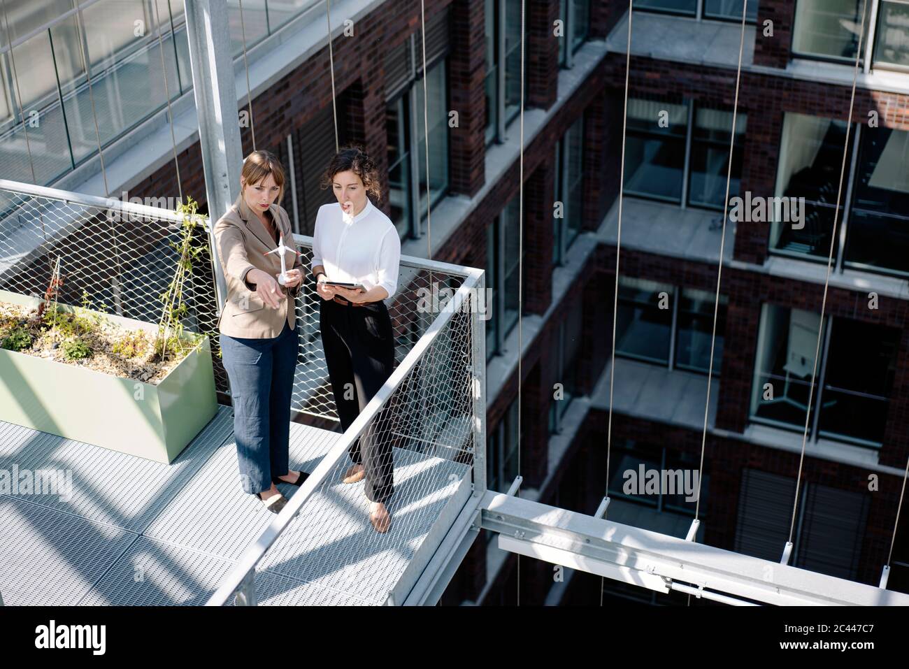 Zwei Geschäftsfrauen mit Tablet und Windturbinenmodell, die auf dem Balkon eines Bürogebäudes eine Besprechung abhalten Stockfoto