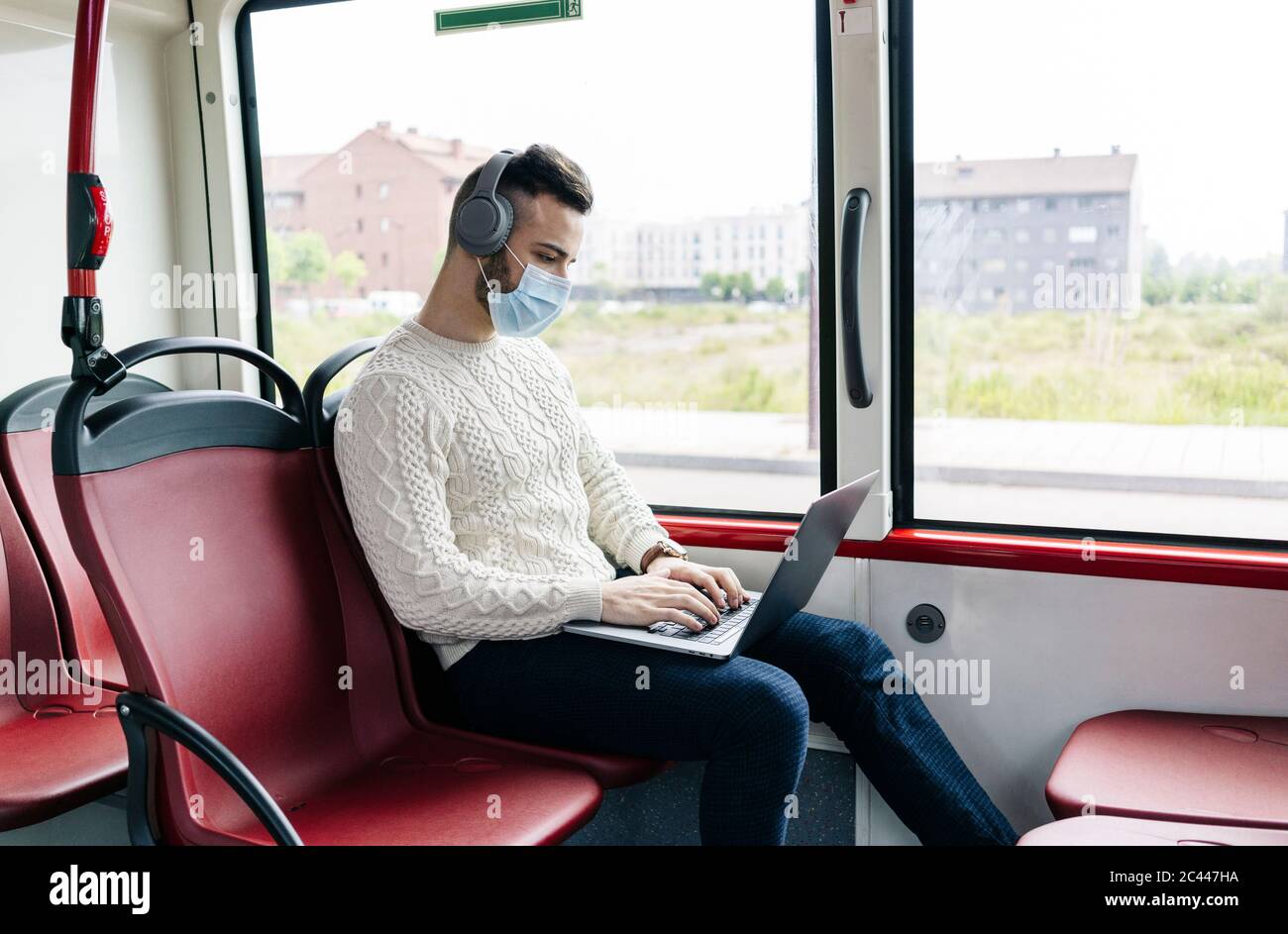 Junger Mann mit Schutzmaske sitzt in öffentlichen Bus mit Kopfhörer und Laptop, Spanien Stockfoto