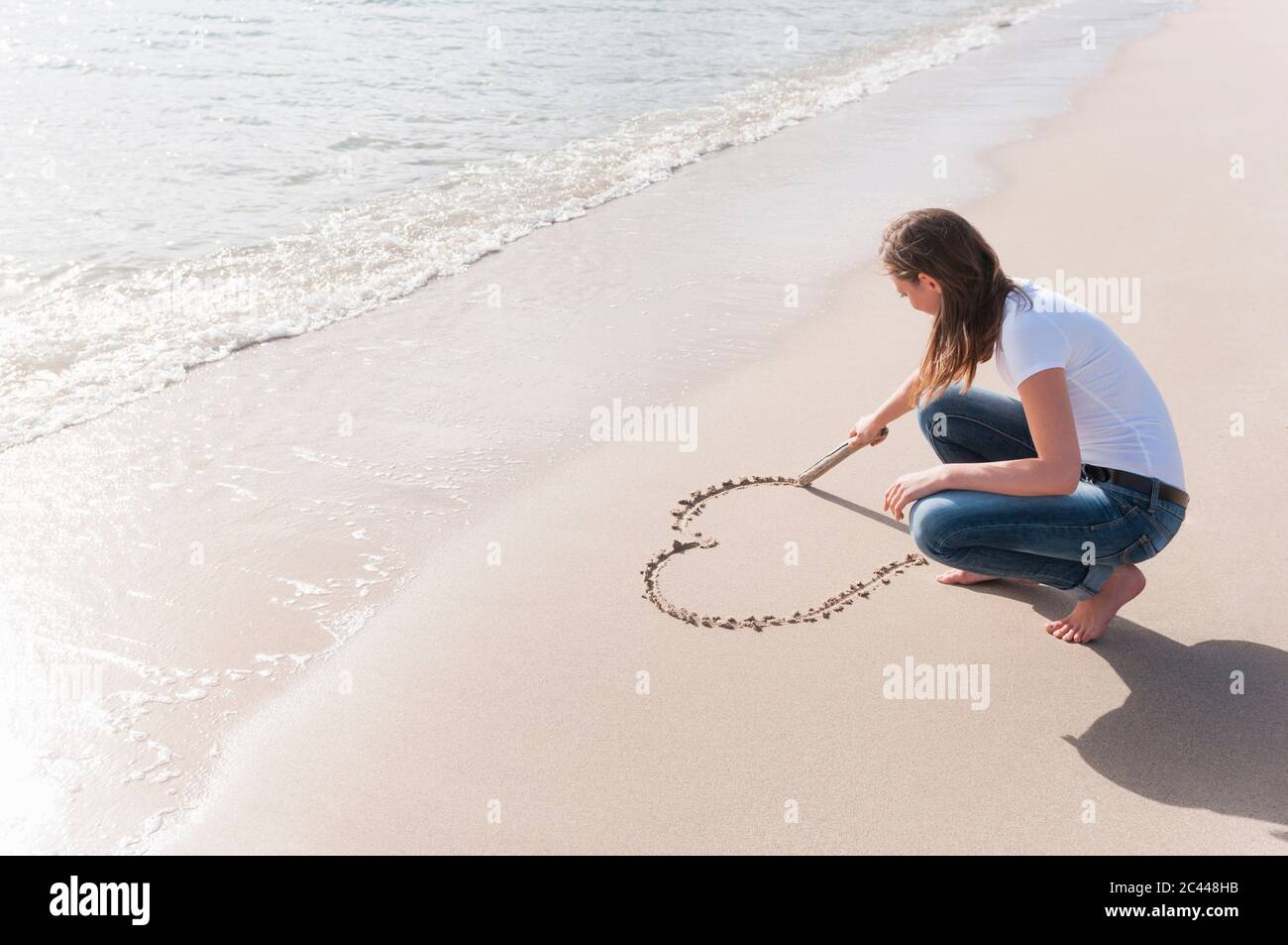 Frau hocken am Strand Kratzen Herz in nassem Sand mit Holzstab, Sardinien, Italien Stockfoto
