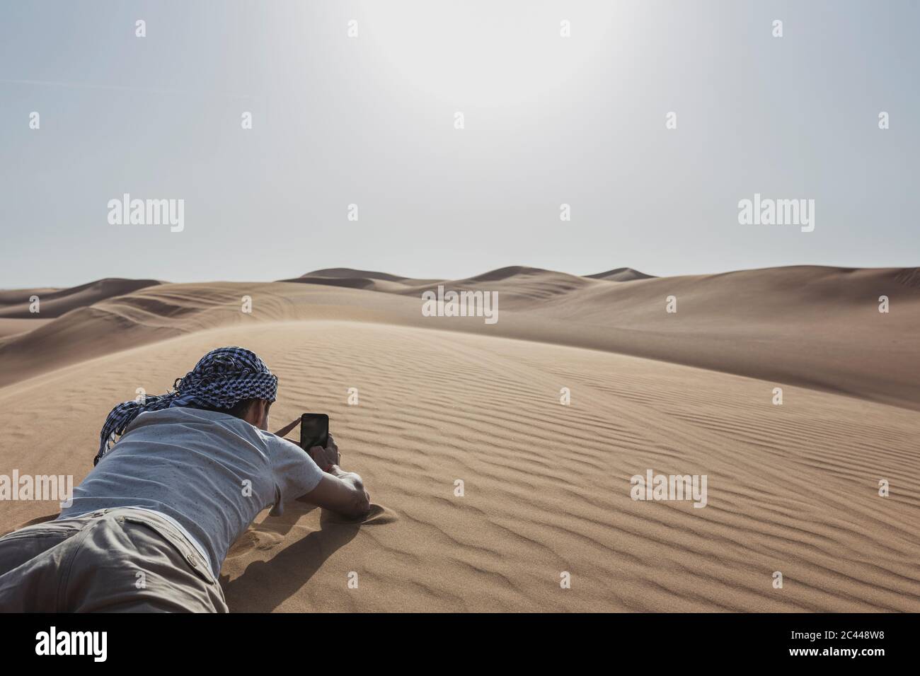 Männlicher Tourist, der mit dem Smartphone fotografiert, während er auf Sanddüne in der Wüste in Dubai, Vereinigte Arabische Emirate, liegt Stockfoto