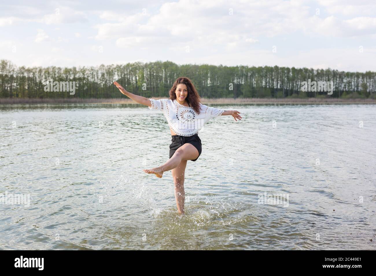Lächelnde junge Frau, die auf einem Bein im See steht und mit Wasser spritzt Stockfoto