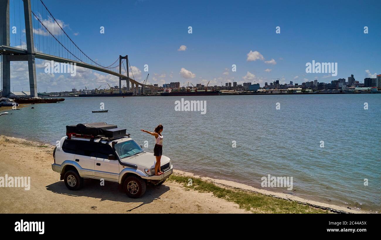 Mosambik, Katembe, Erwachsene Frau mit erhobenen Armen auf Stoßstange von 4x4 Auto bewundern Blick auf Maputo Bay mit Stadt und Maputo-Katembe Brücke im Hintergrund Stockfoto